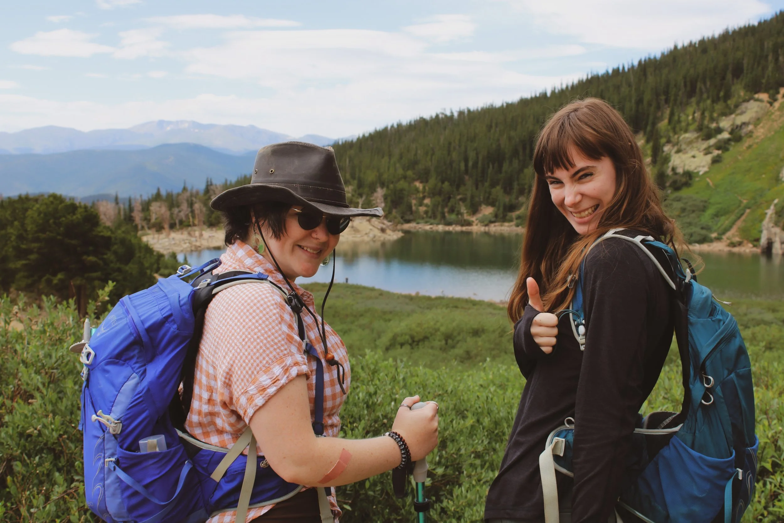Two smiling women with backpacks hiking near a lake surrounded by mountains and green trees.