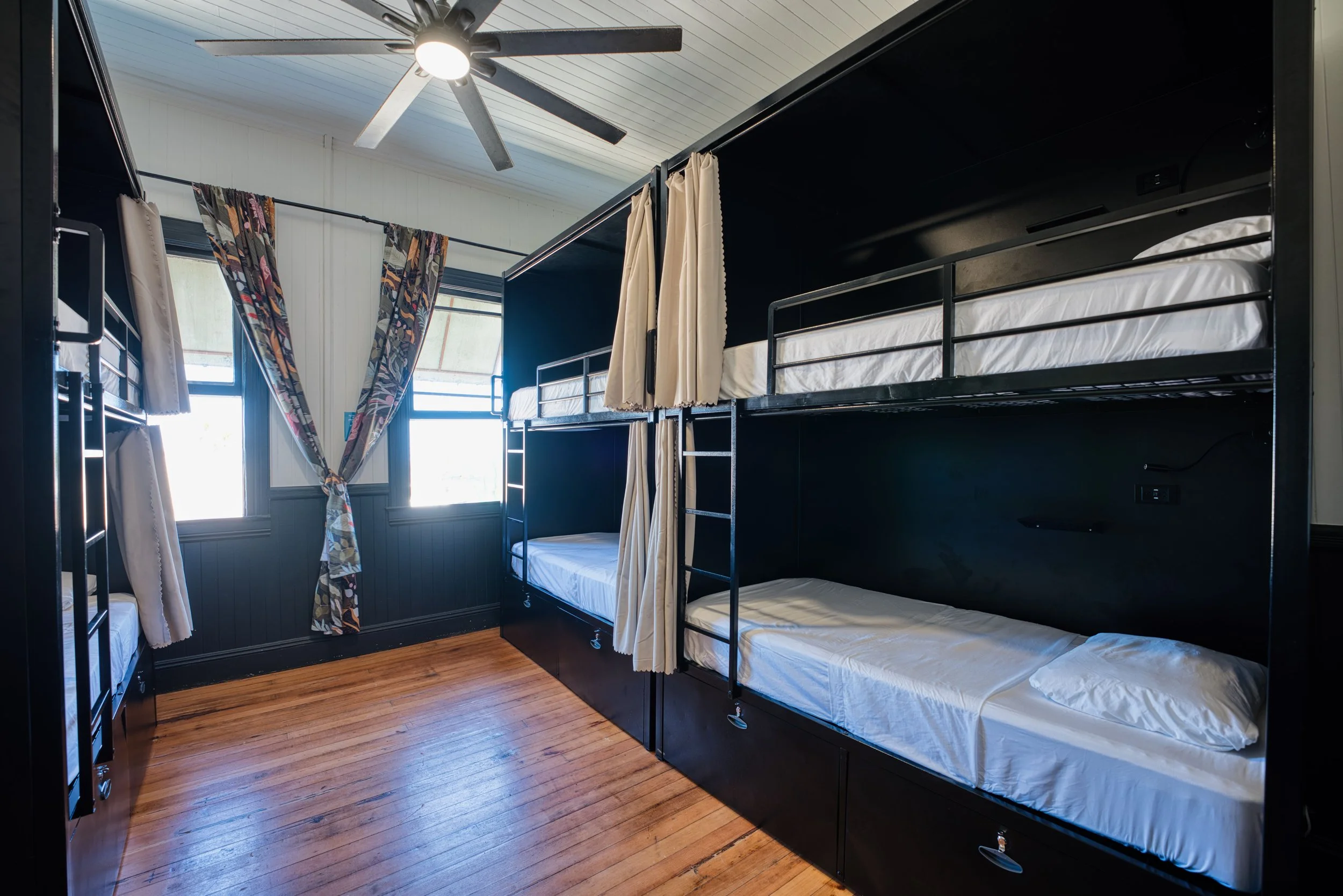 Room with two sets of bunk beds, black metal frames, white bedding, curtains, two windows with patterned curtains, wooden floor, ceiling fan with light.