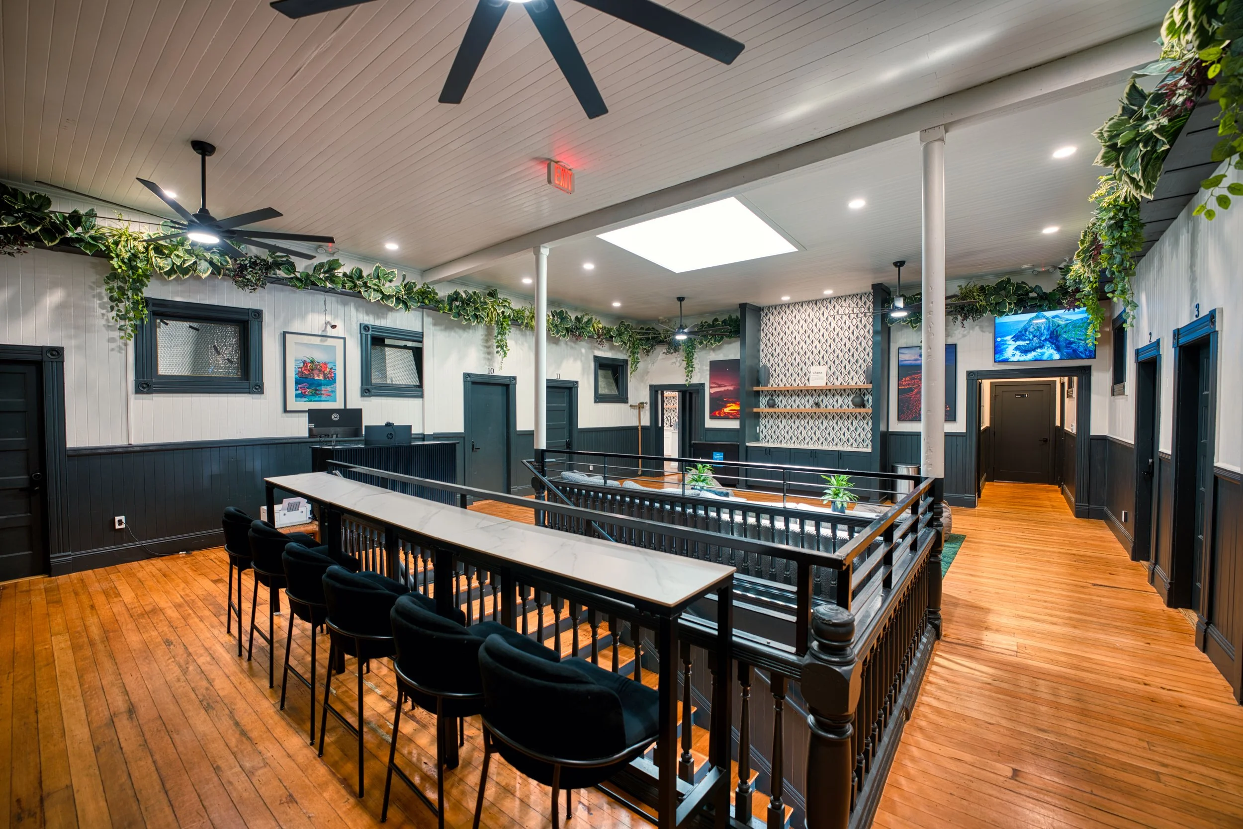 Interior of a well-lit cafe with wooden floors, black wainscoting, green plants, wall art, a marble-topped counter, and television screens.