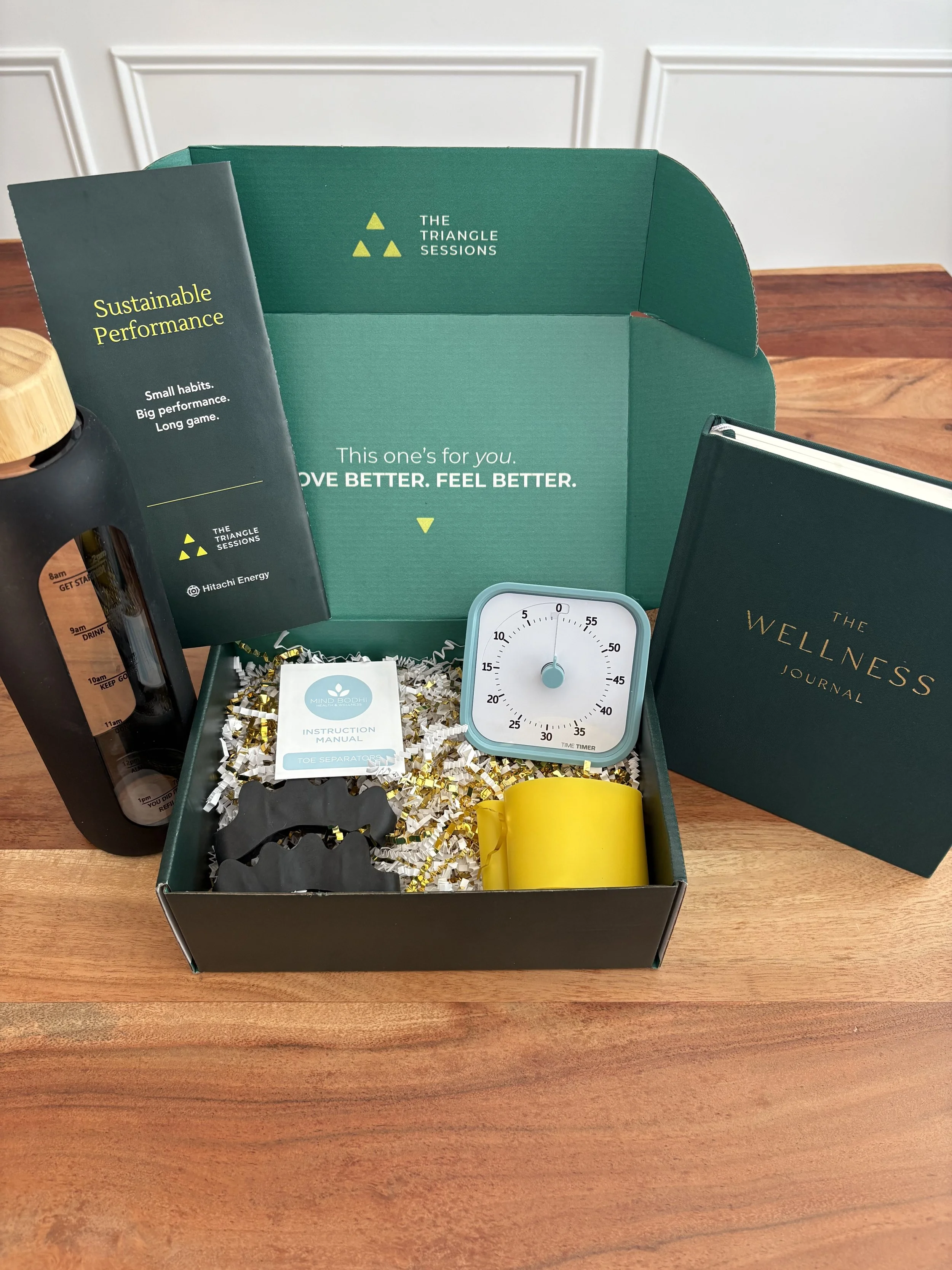 Various wellness and productivity items in a green box on a wooden table, including a book titled "The Wellness Journal", a timer, a booklet titled "Sustainable Performance", a small instruction manual, a yellow clip, a toe separator, a black bottle, and a black spiral notebook.