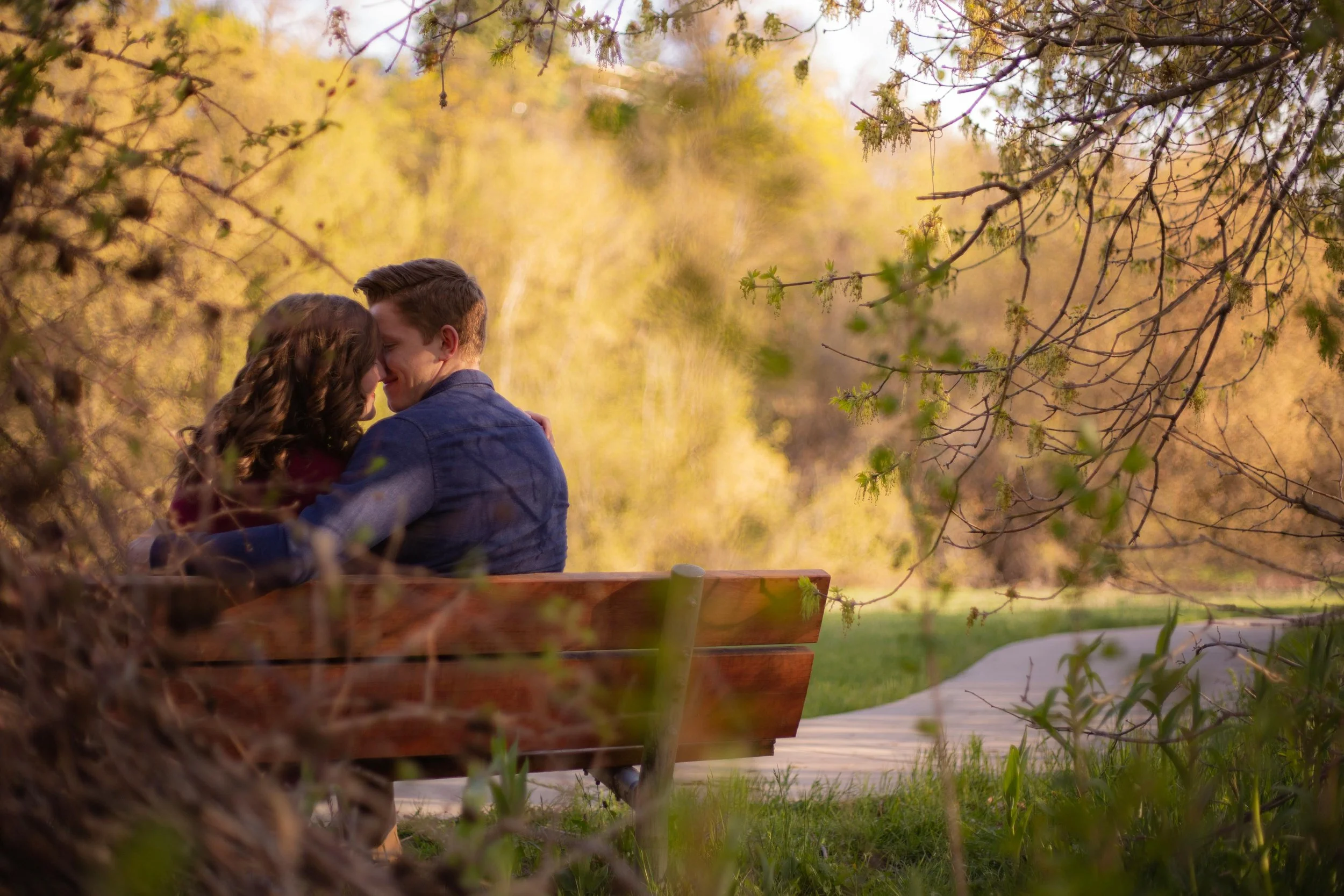A couple sitting close together on a park bench, surrounded by trees and greenery, sharing an intimate moment in a peaceful outdoor setting during daytime.