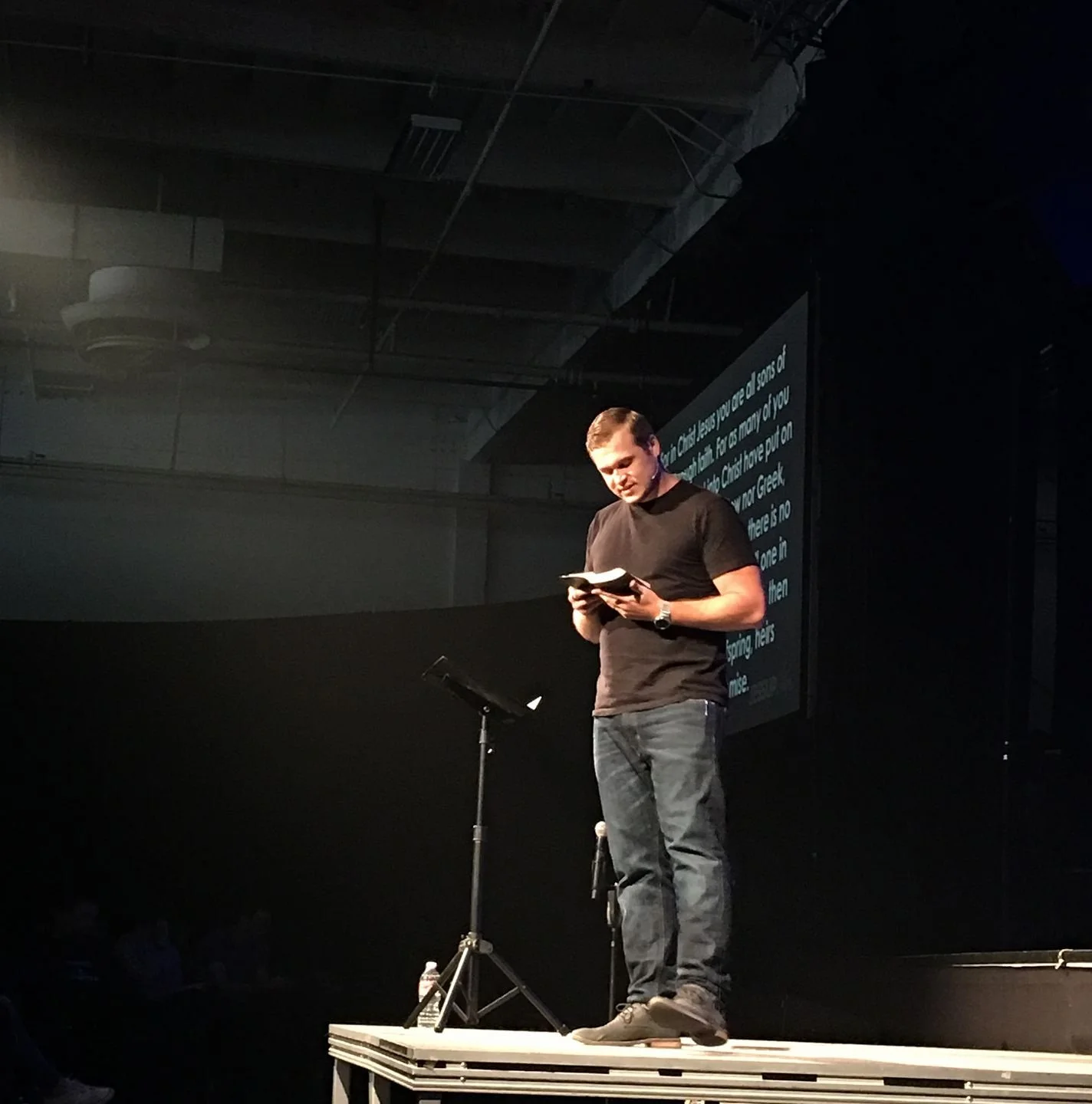 A man in a black t-shirt and jeans standing on a stage, reading from a book, with a microphone stand and a large screen displaying text behind him.