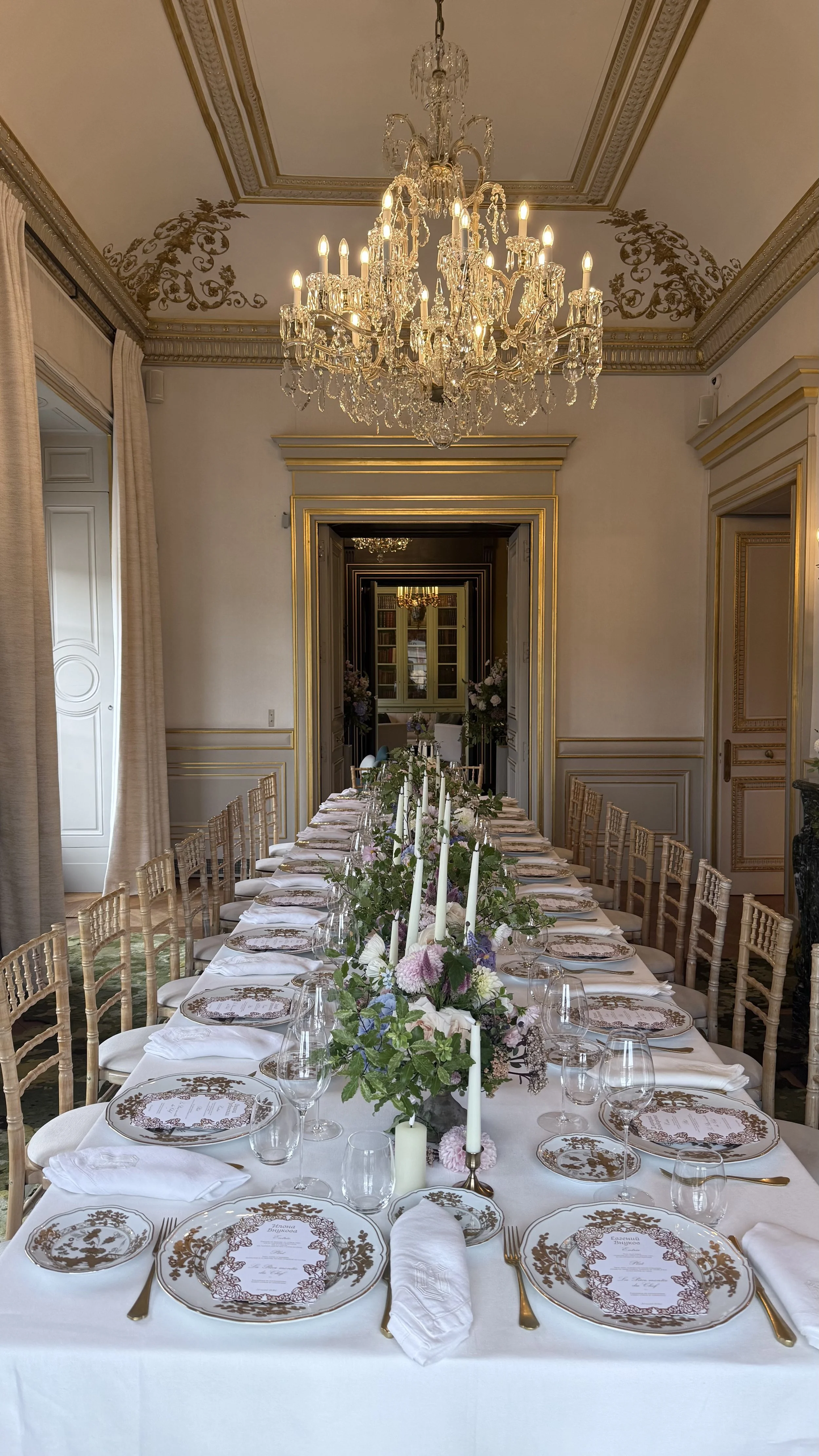 Une grande table dressée pour un repas ou un événement, ornée de vaisselle élégante, de fleurs, de bougies et de couvre-plat, dans une salle richement décorée avec un chandelier en cristal suspendu au plafond.