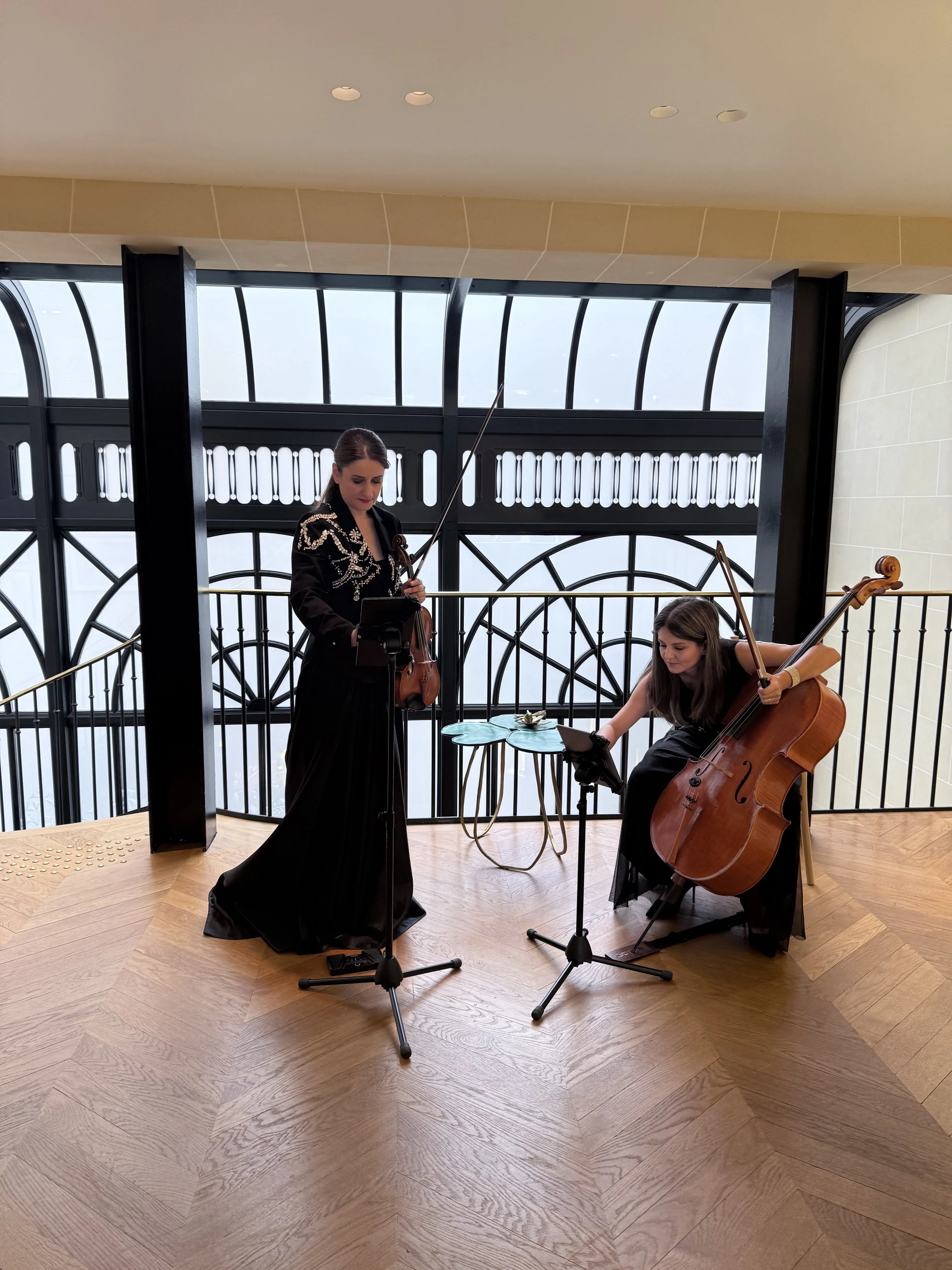 Deux femmes jouant de la musique dans une salle avec un grand vitrage en arrière-plan, l'une avec un violon et l'autre avec un violoncelle, toutes deux regardant des écrans ou des feuilles de musique.