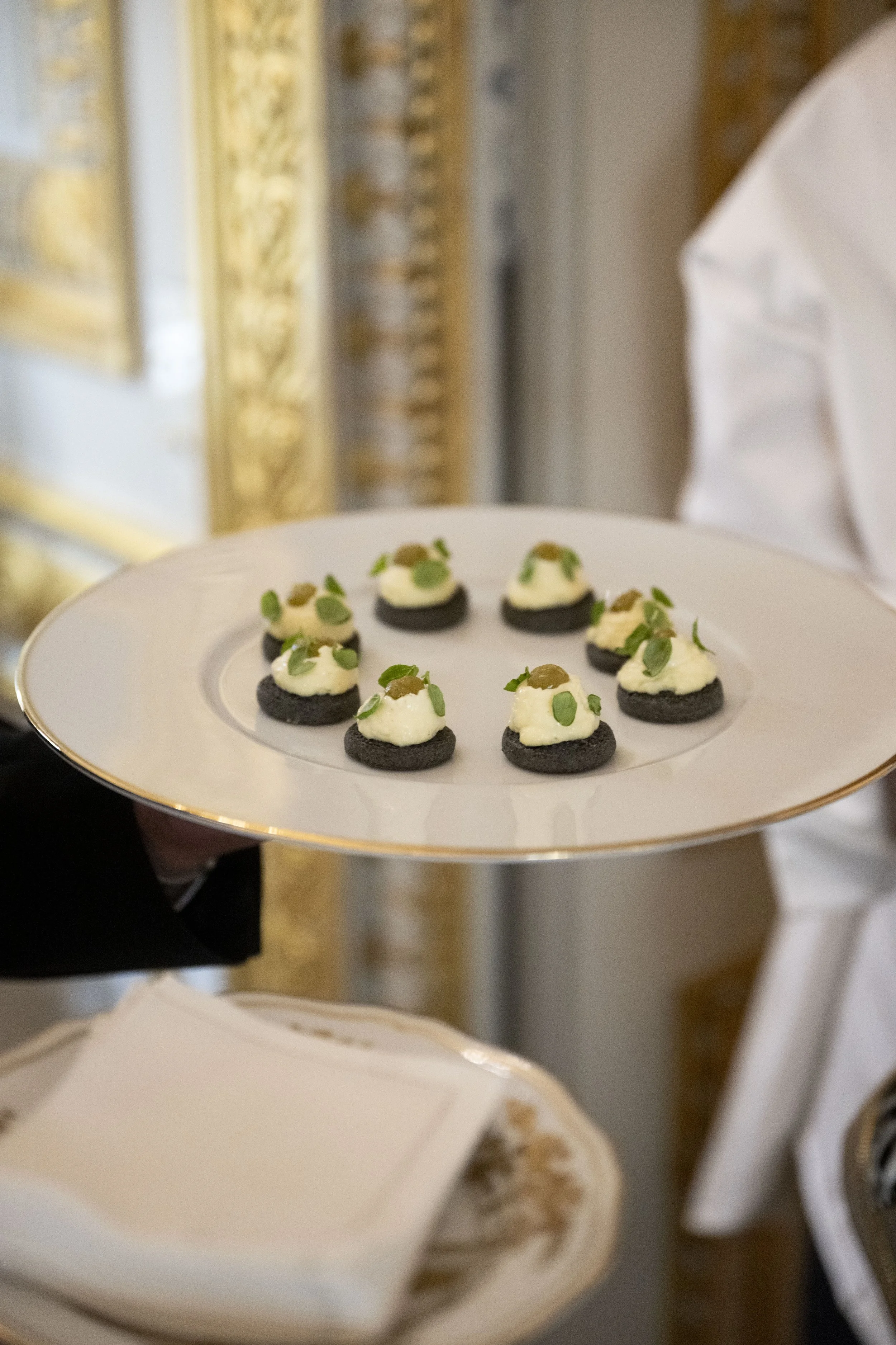Petites bouchées servies sur une assiette blanche avec une bordure dorée, garnies de crème, de petits légumes verts, et de moultes conservés, présentées dans un décor élégant.