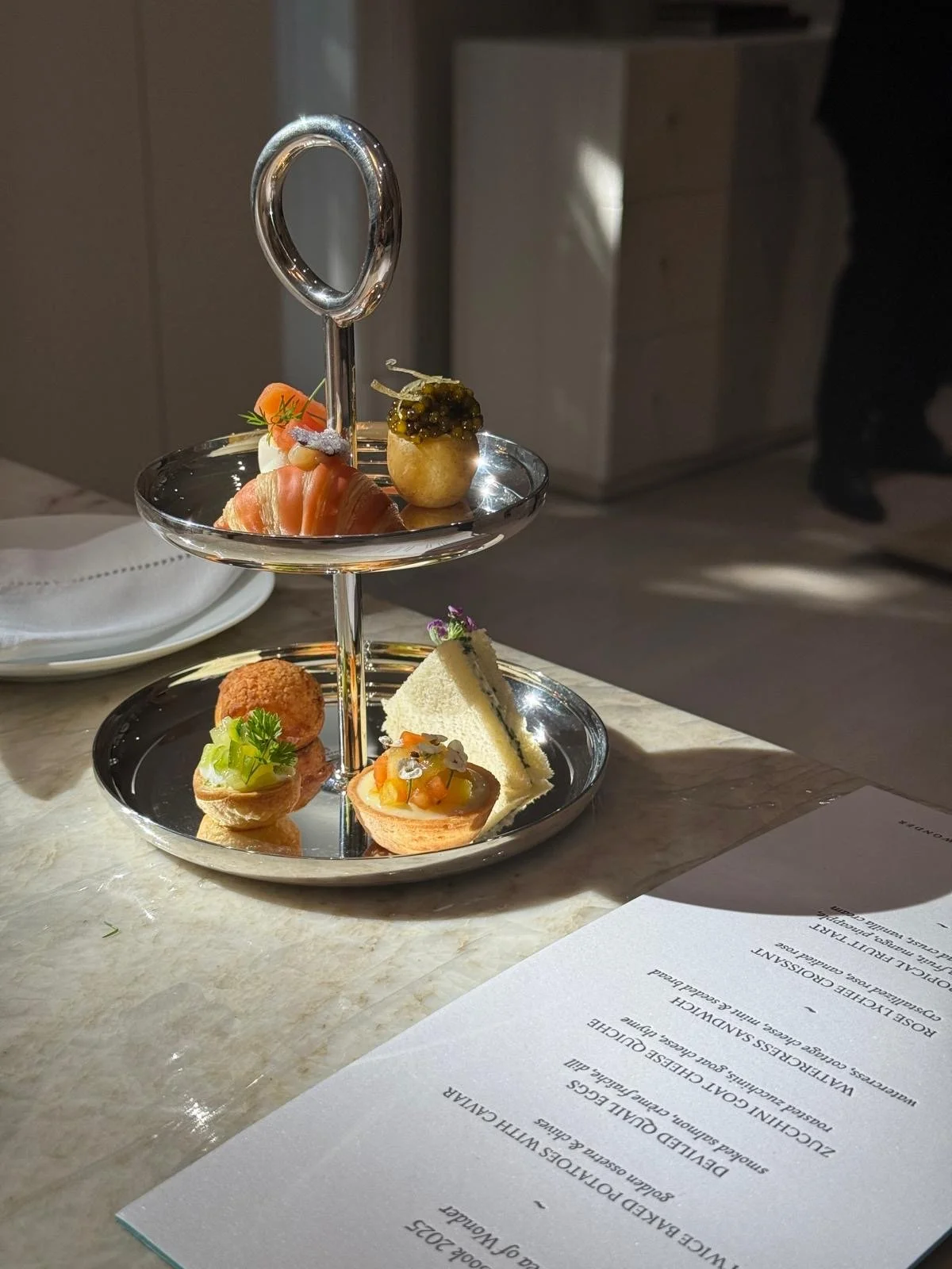 Plateau de dégustation avec plusieurs petites portions de nourriture sur un support à plusieurs niveaux, éclairé par la lumière naturelle, avec une liste de menu partiellement visible à côté.