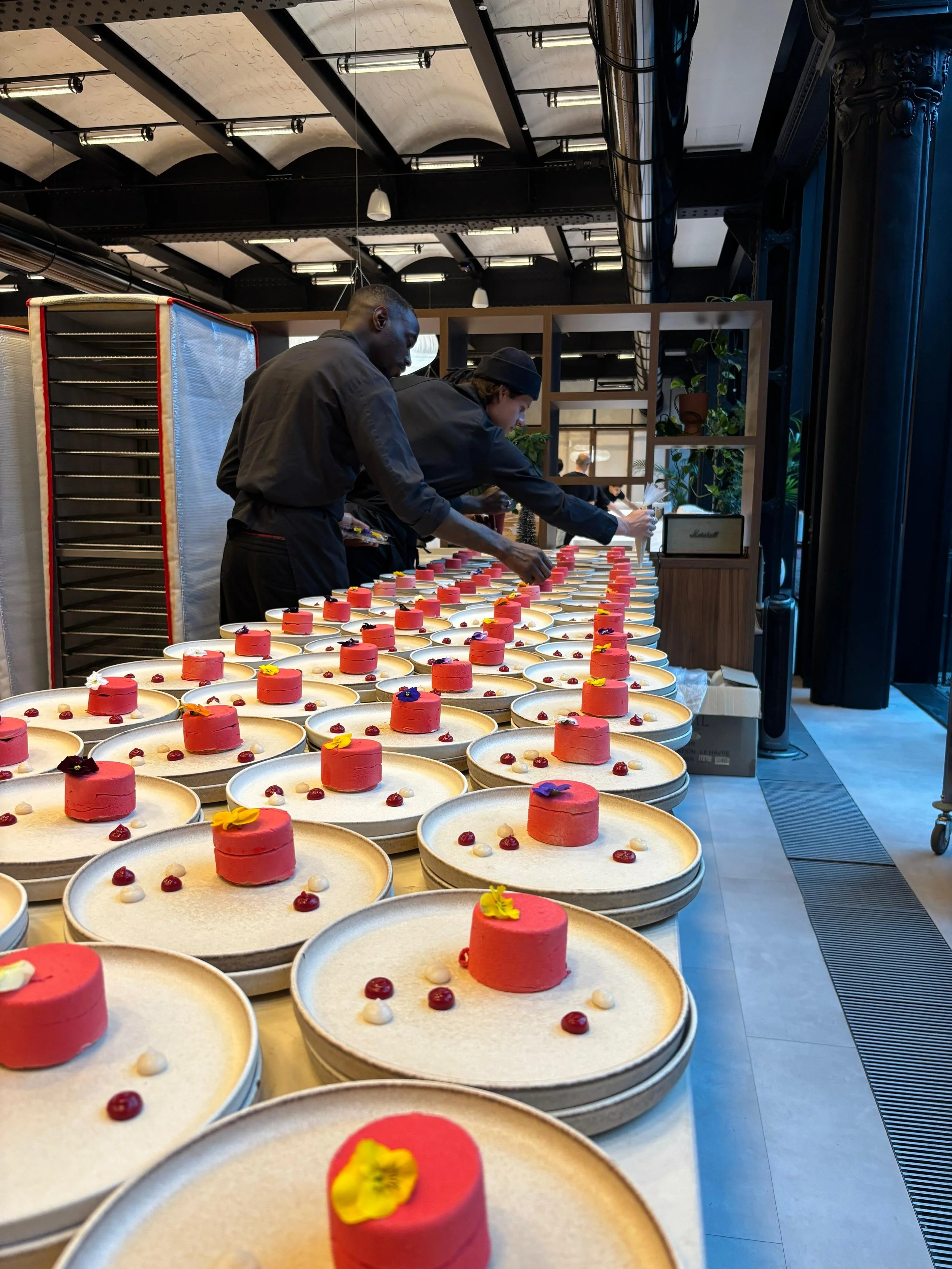 Plusieurs chefs en noir préparant des desserts modernes sur une longue table avec de nombreux gâteaux ronds rose et décorés de fleurs et de petits fruits dans un espace industriel.