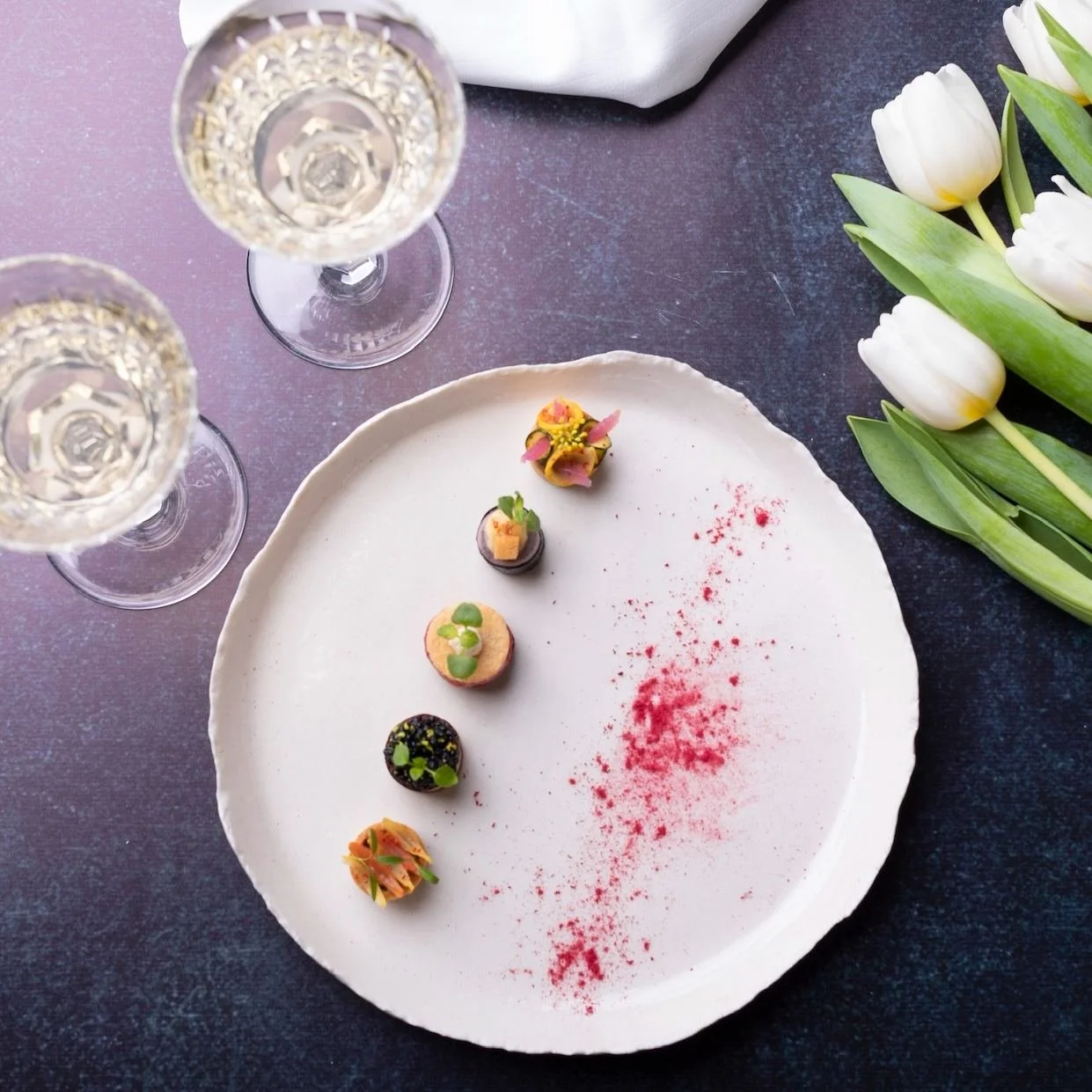 Assiette élégante avec six petits desserts colorés, décorée de poudre rouge, à côté de tulipes blanches, deux verres de champagne ou vin pétillant, sur une table sombre.