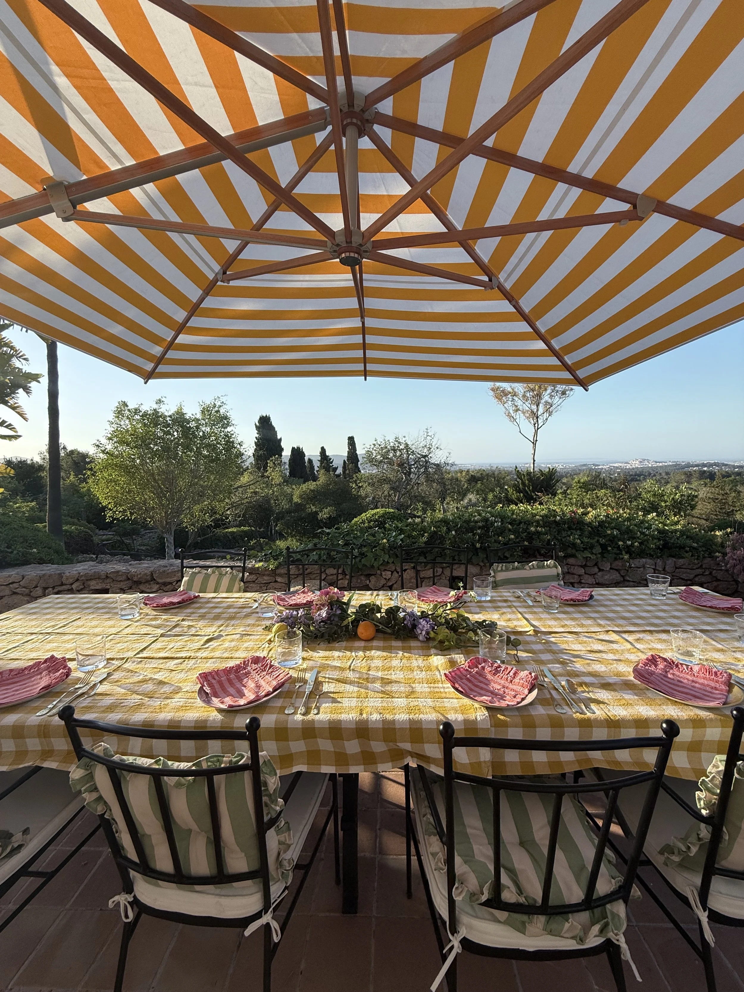 Table de repas en plein air sous un grand parasol rayé jaune et blanc, avec un décor de verdure et de paysages en arrière-plan.