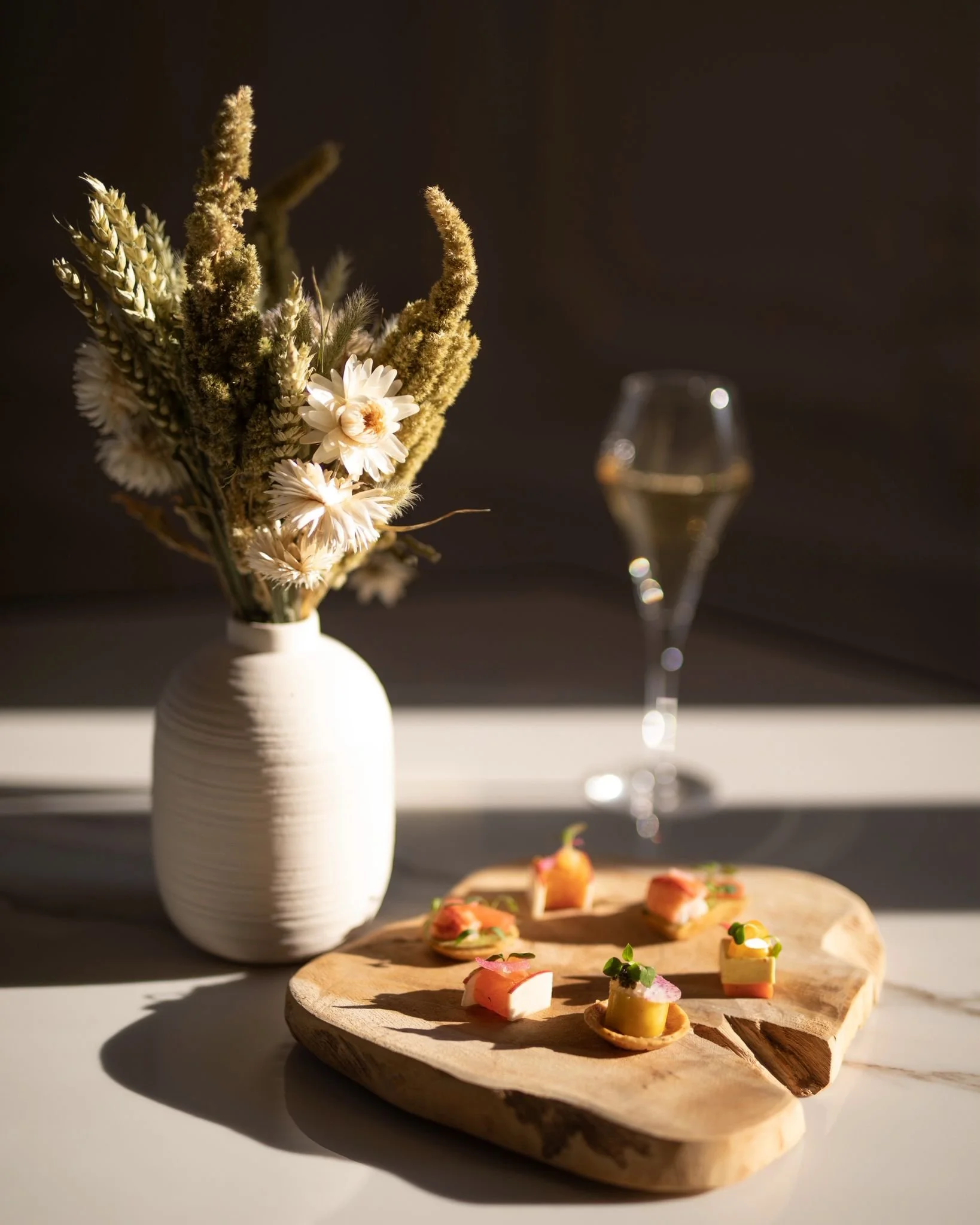 Un vase blanc avec des fleurs séchées et un plateau en bois avec des amuse-bouches, une coupe de champagne à l'arrière