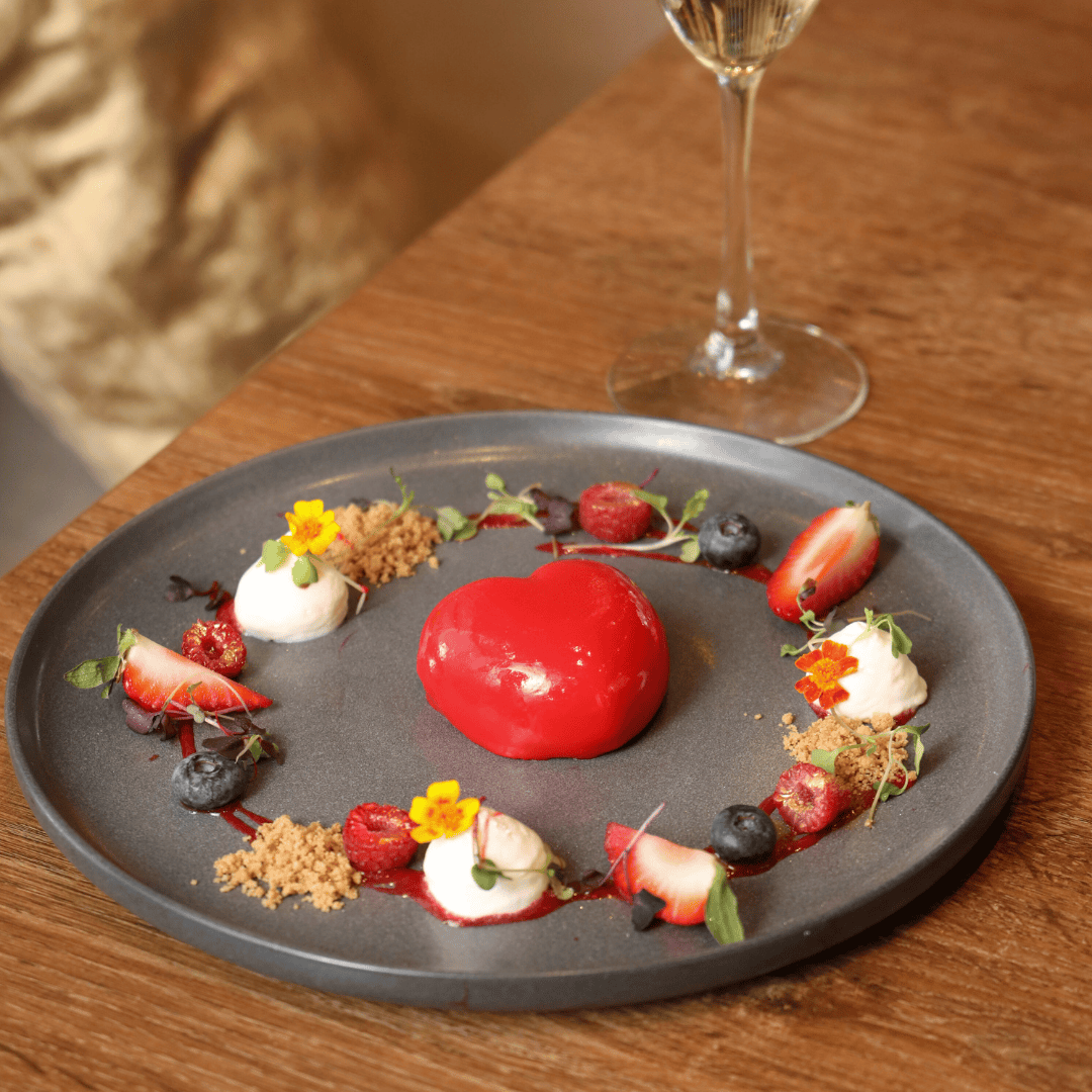 Elegant dessert plate with a glossy red domed element in the center, surrounded by strawberries, blueberries, dollops of cream, edible flowers, and crumbs on a dark grey plate, next to a glass of white wine on a wooden table.