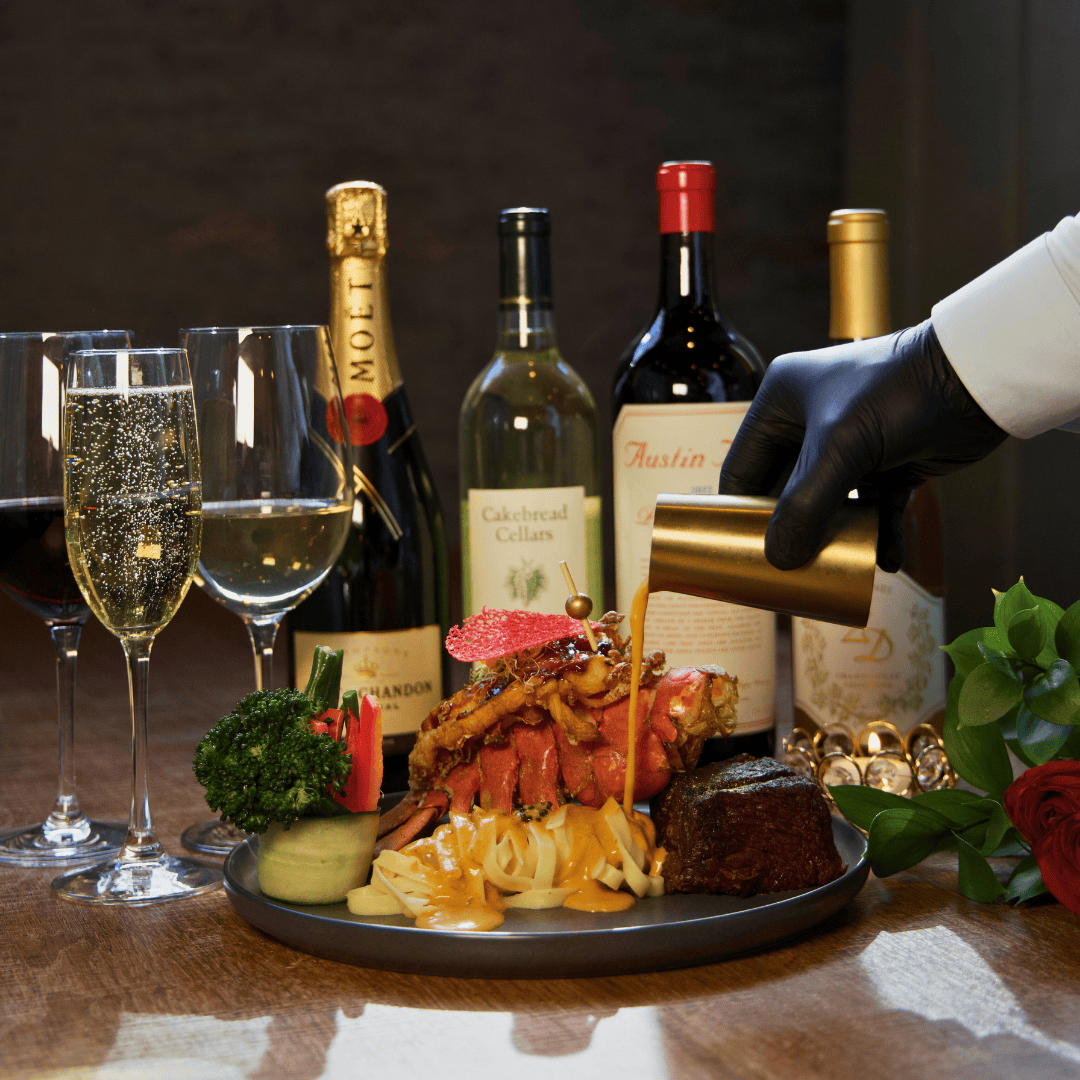 A gourmet steak dinner being poured with sauce, with wine bottles, glasses of champagne and red wine in the background, and a bouquet of flowers on the side.