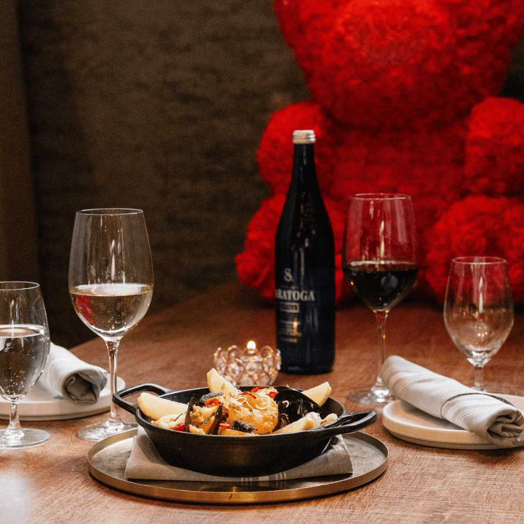 A restaurant table set with three wine glasses, one of which has white wine and another with red wine, a bottle of wine, a small cast iron skillet with a seafood dish topped with lemon wedges, and neatly rolled napkins on white plates.