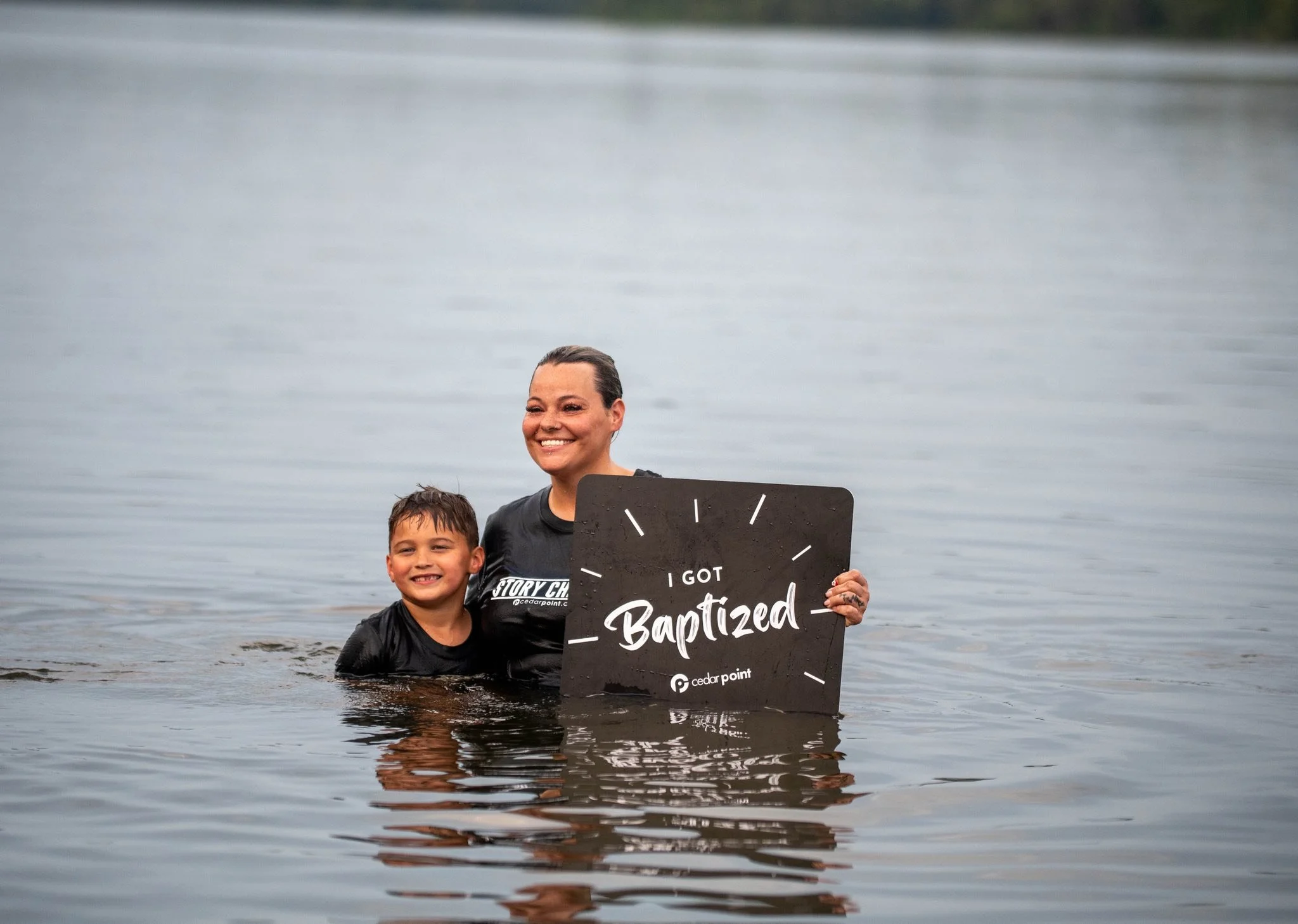 A woman and a boy are in a body of water, smiling, holding a sign that says 'I got baptized' with a logo and the words 'cedar point' on it.