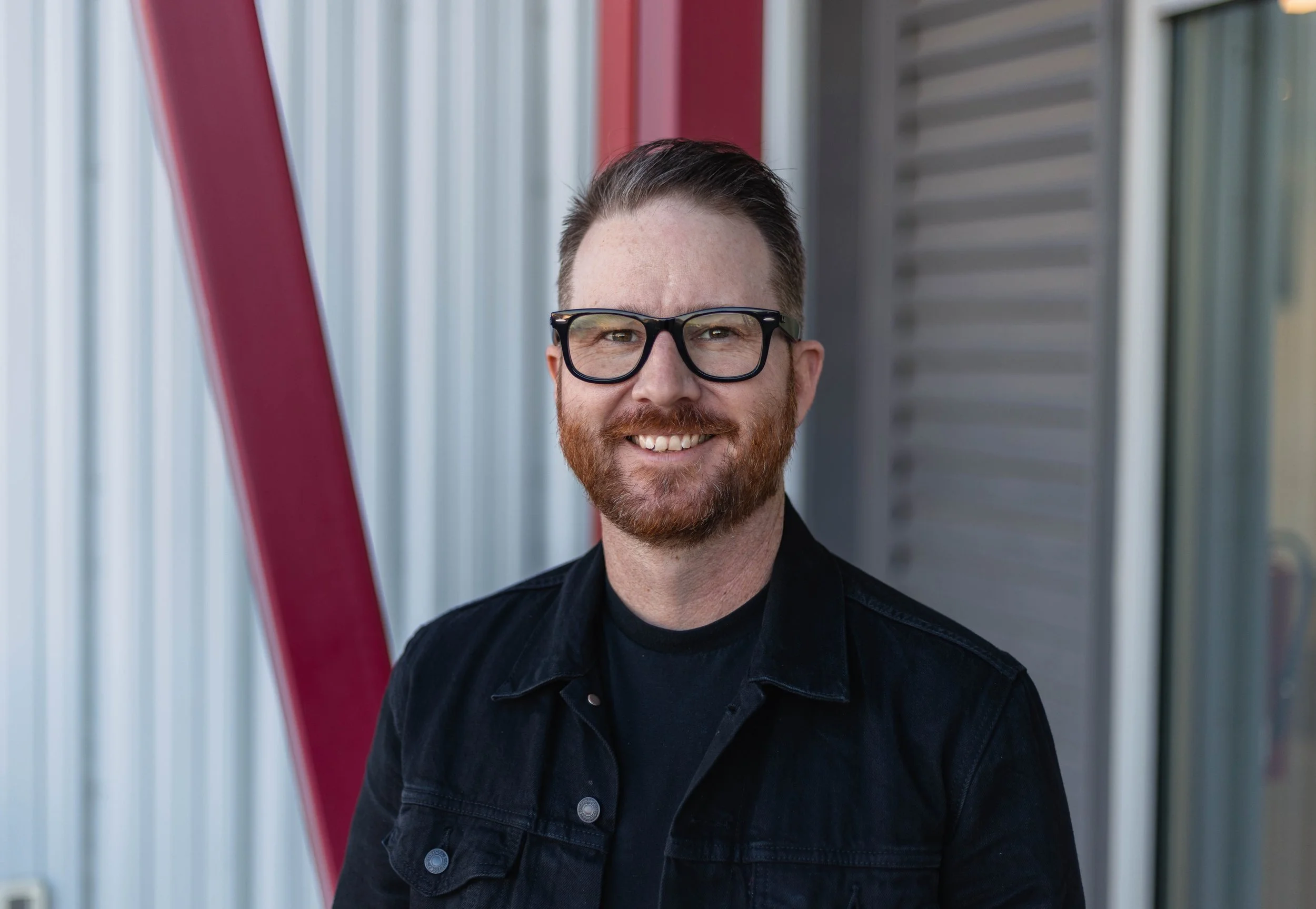 A man with glasses and a beard standing outdoors in front of a building with red and blue siding, looking at the camera.