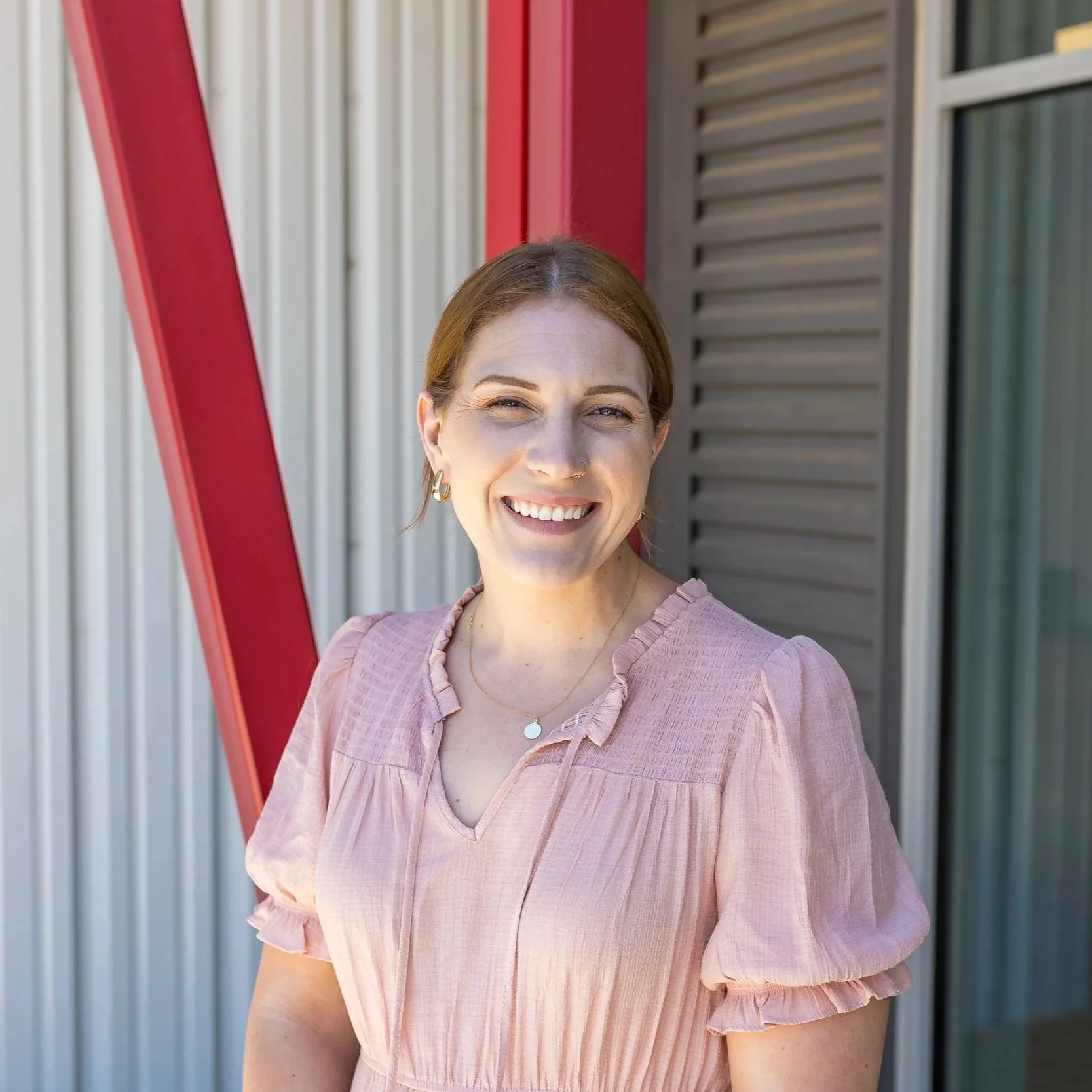 A smiling woman with shoulder-length red hair wearing a light green dress standing outside near a building with glass windows.