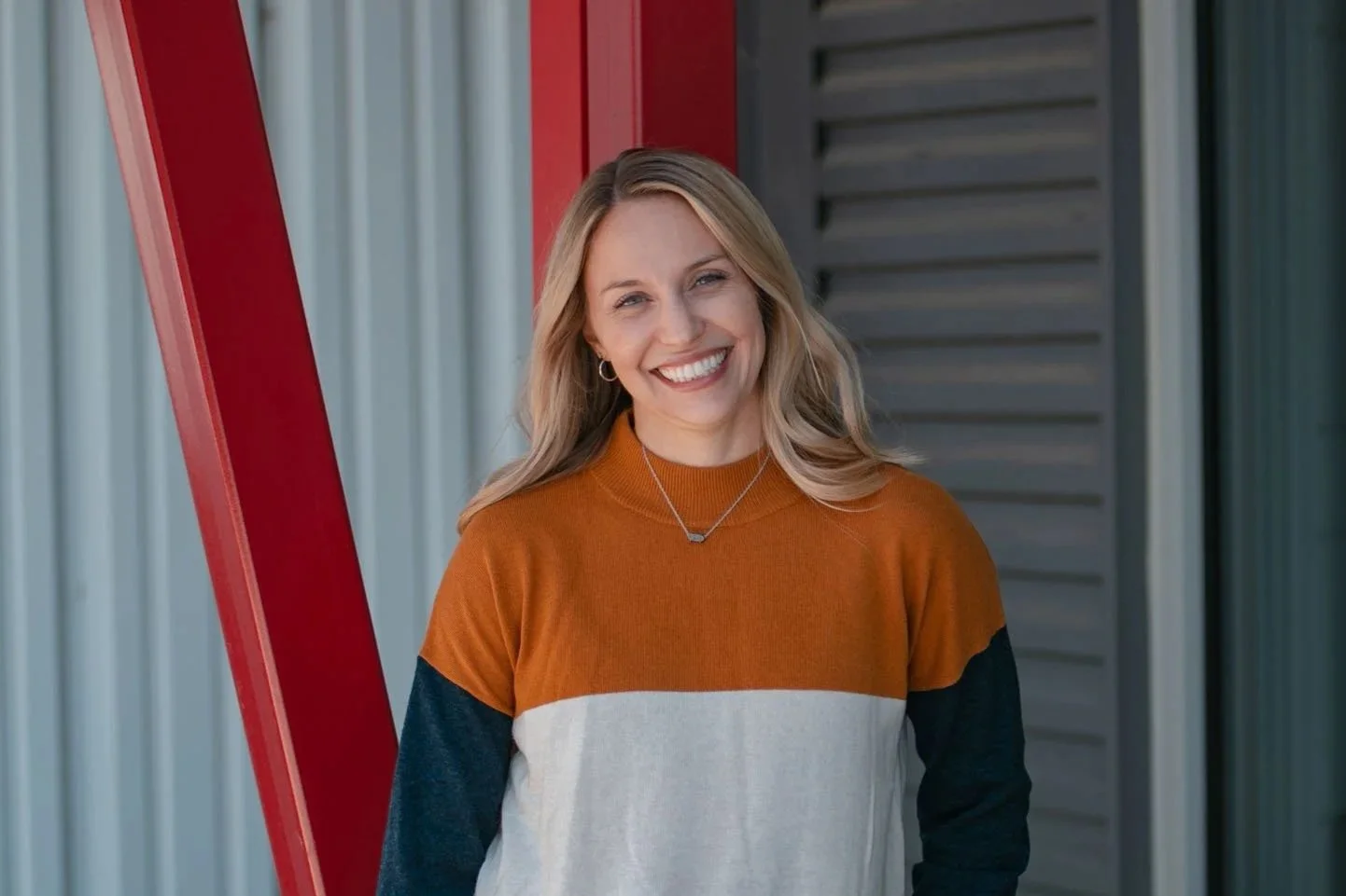A smiling woman with blonde hair wearing a color-blocked long sleeve shirt, standing outdoors next to metal siding and a red structure.