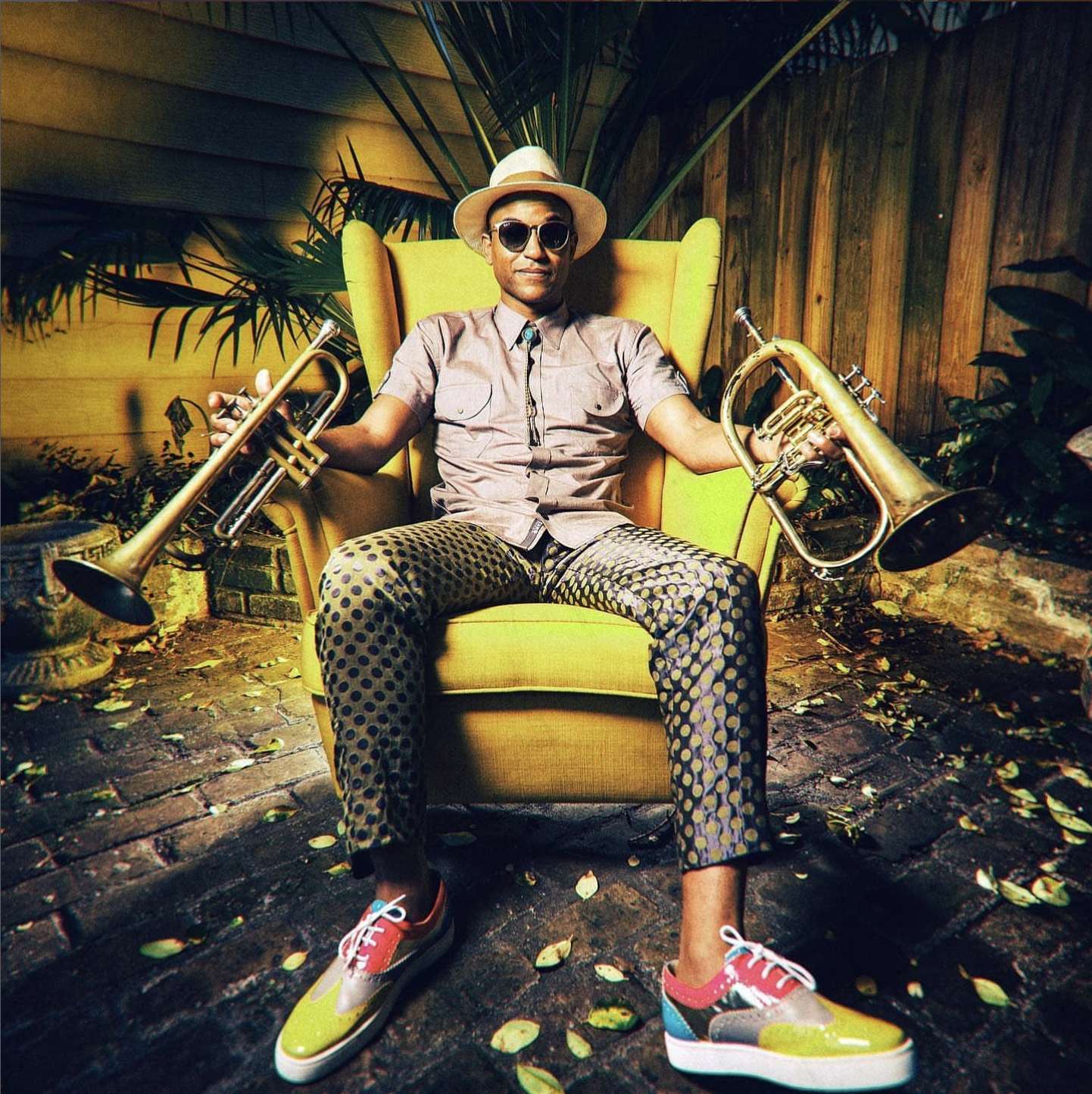 A man sitting in a yellow armchair outdoors, holding brass trumpet and cornet. He wears a beige fedora, sunglasses, a short-sleeve button-up shirt, patterned pants, and colorful sneakers. There are plants and a wooden fence in the background and leaves on the ground.