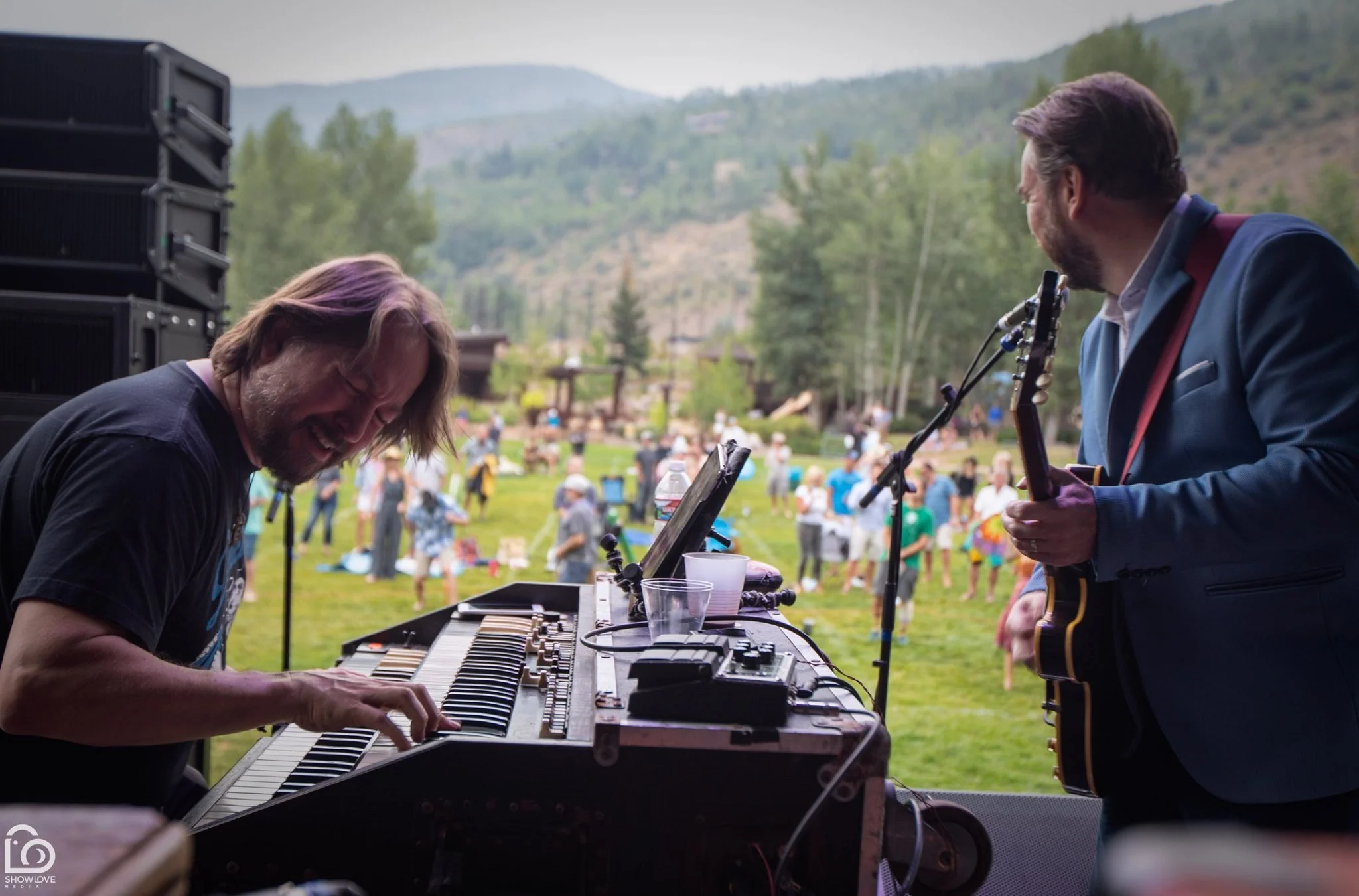 Musicians performing outdoors at a festival, one playing keyboard and the other playing guitar, with a crowd and mountains in the background.