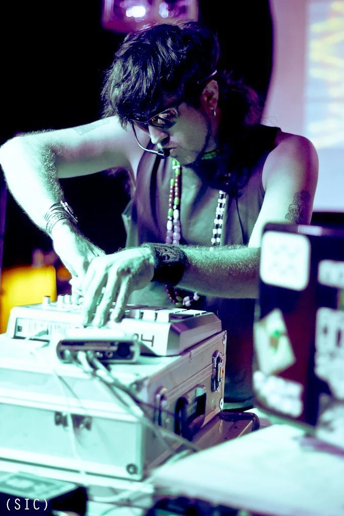 A person with dark hair, glasses, and tattoos adjusts a DJ mixer, wearing a headset, in a dimly lit environment with colorful lights.