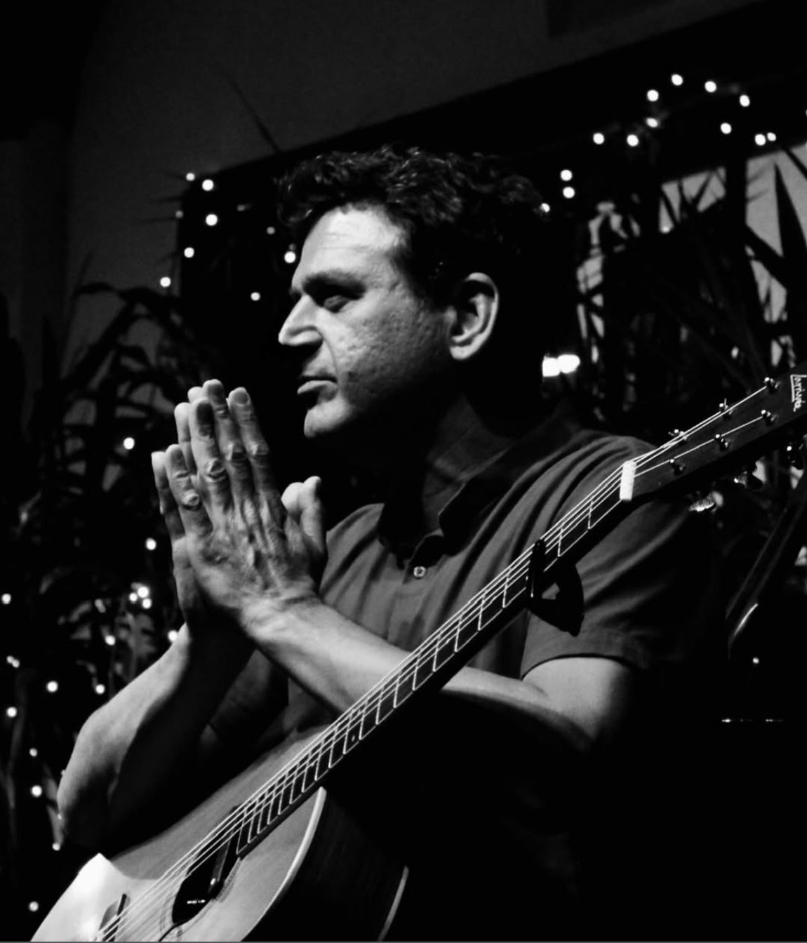 A man with dark hair and a guitar, seen in profile with closed eyes, hands pressed together in prayer, in a dimly lit setting with decorative lights and plants in the background.