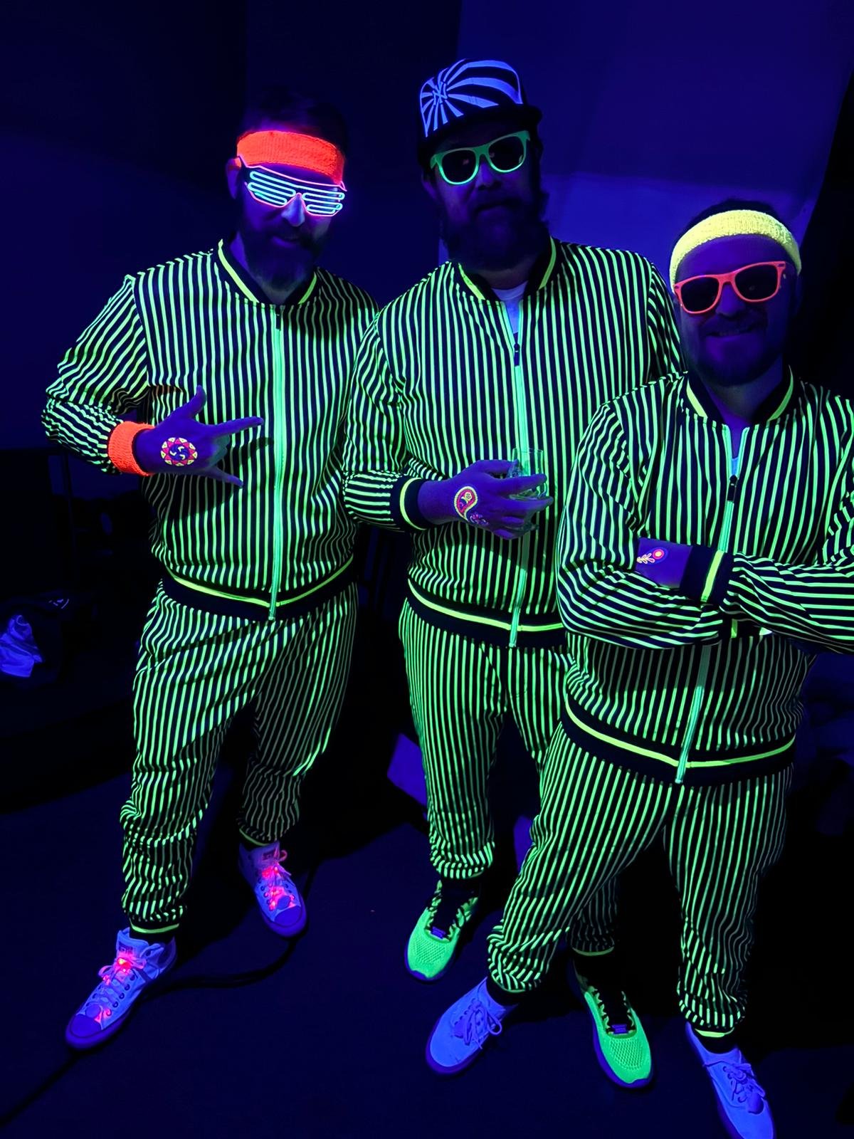 Three men wearing glowing blacklight-striped outfits with matching shoes and neon accessories, posing with neon glasses and caps in a dark room with blacklights.