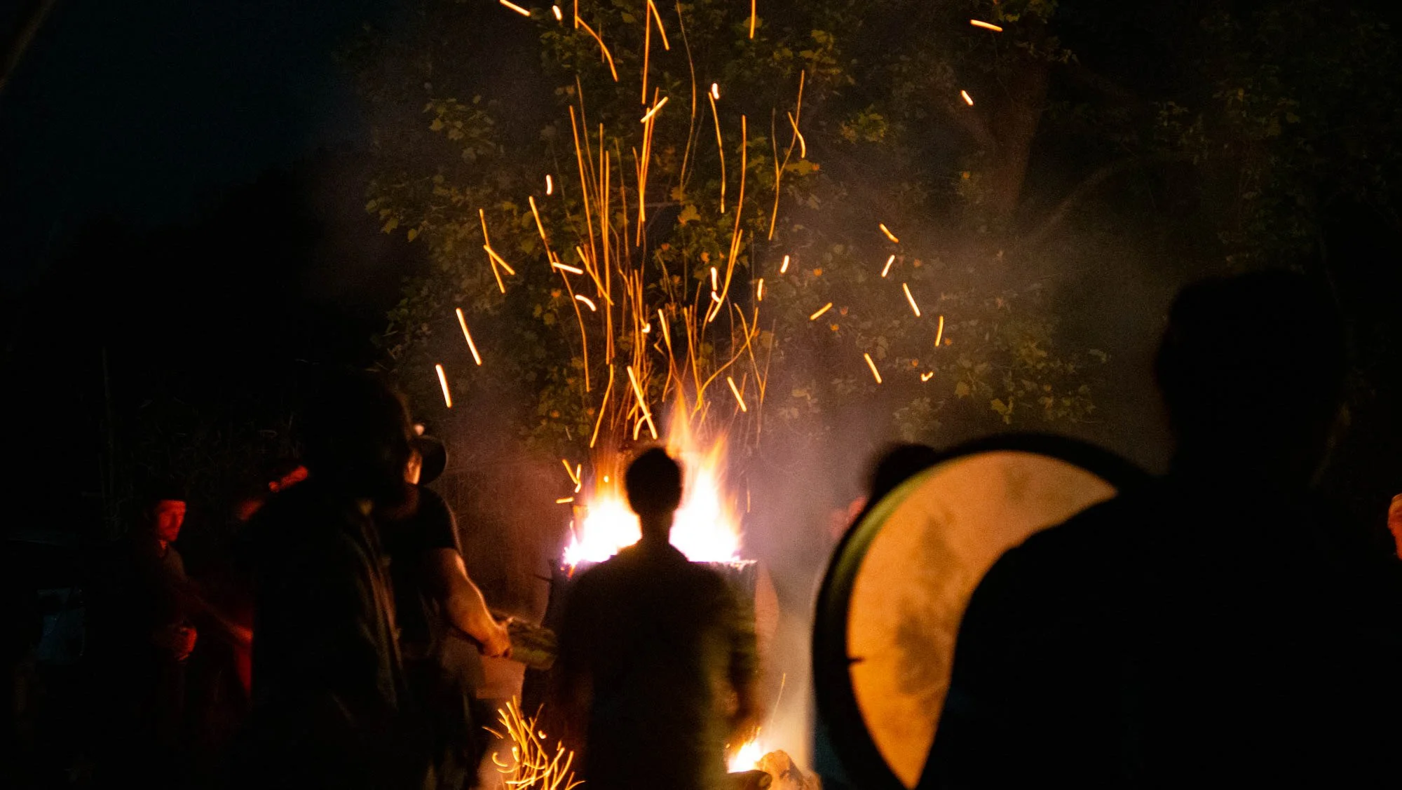 Nighttime outdoor scene with people gathered around a bonfire, sparks flying into the air, and trees in the background.