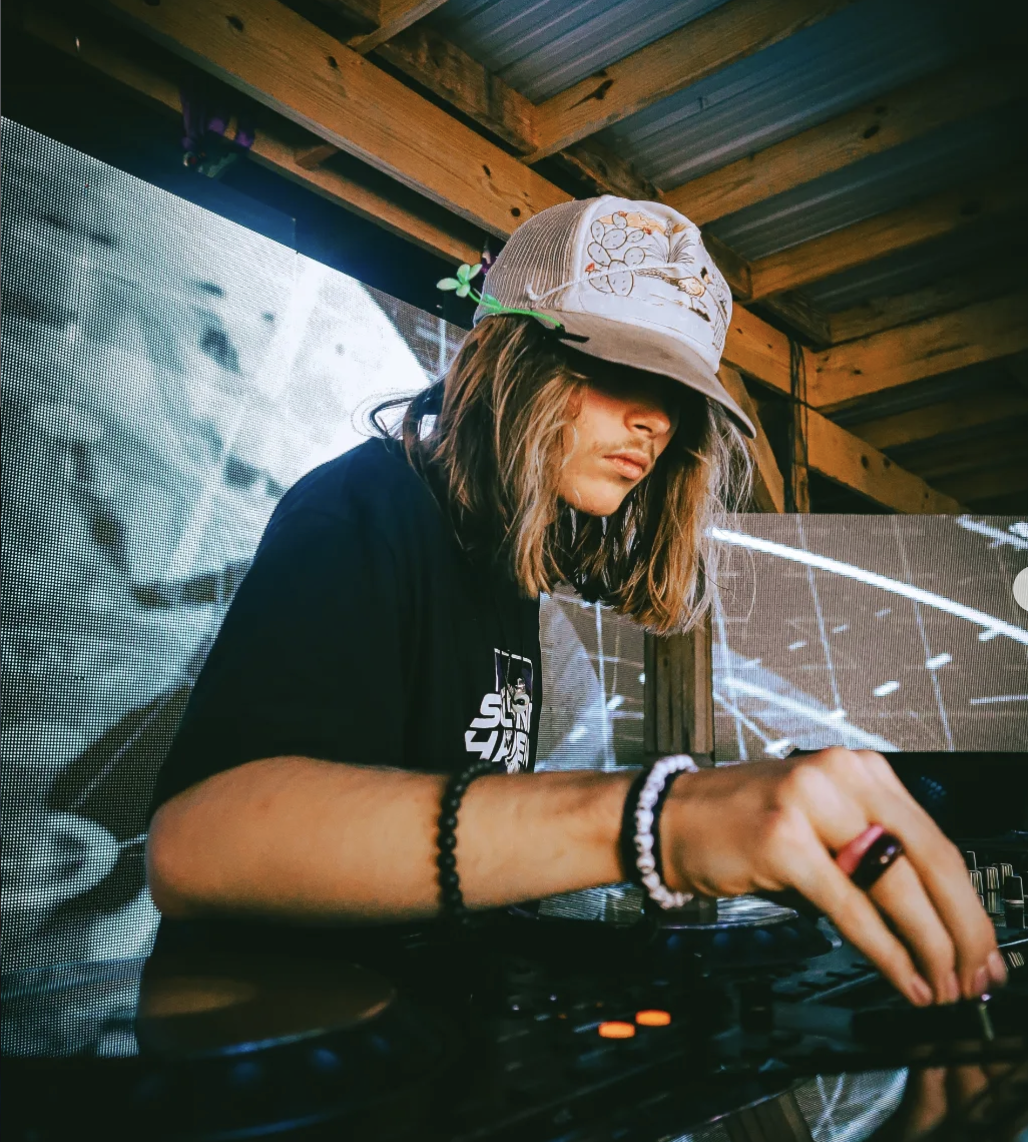 A young DJ performing at an outdoor event, wearing a beige cap with floral embroidery, black t-shirt, and bracelets on her left wrist, with a DJ console in front of him.