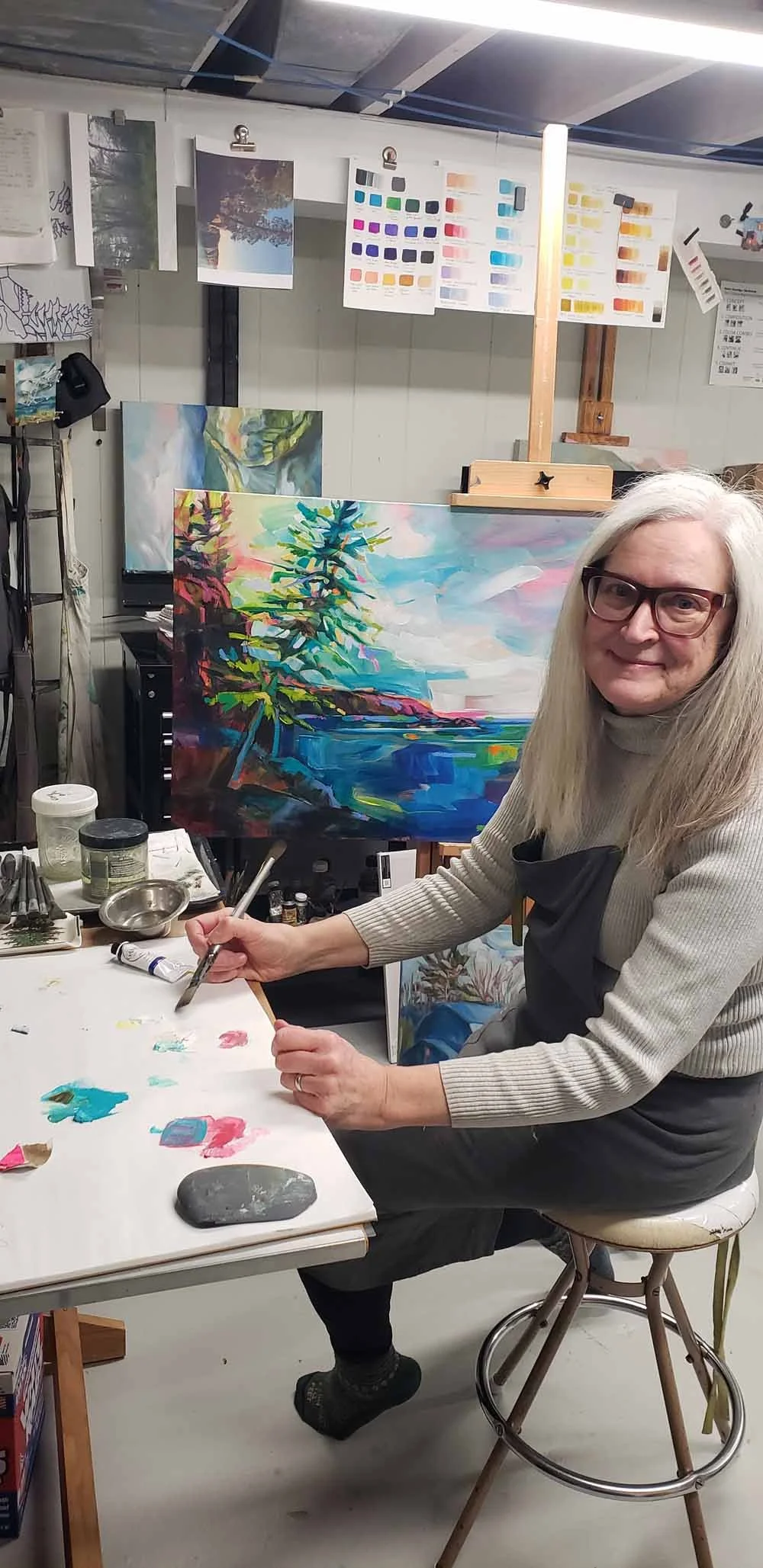 A woman with glasses and long gray hair sitting at a table in an art studio, painting a colorful landscape on a canvas with a tree, hills, and sky in the background, surrounded by painting supplies and other artwork.