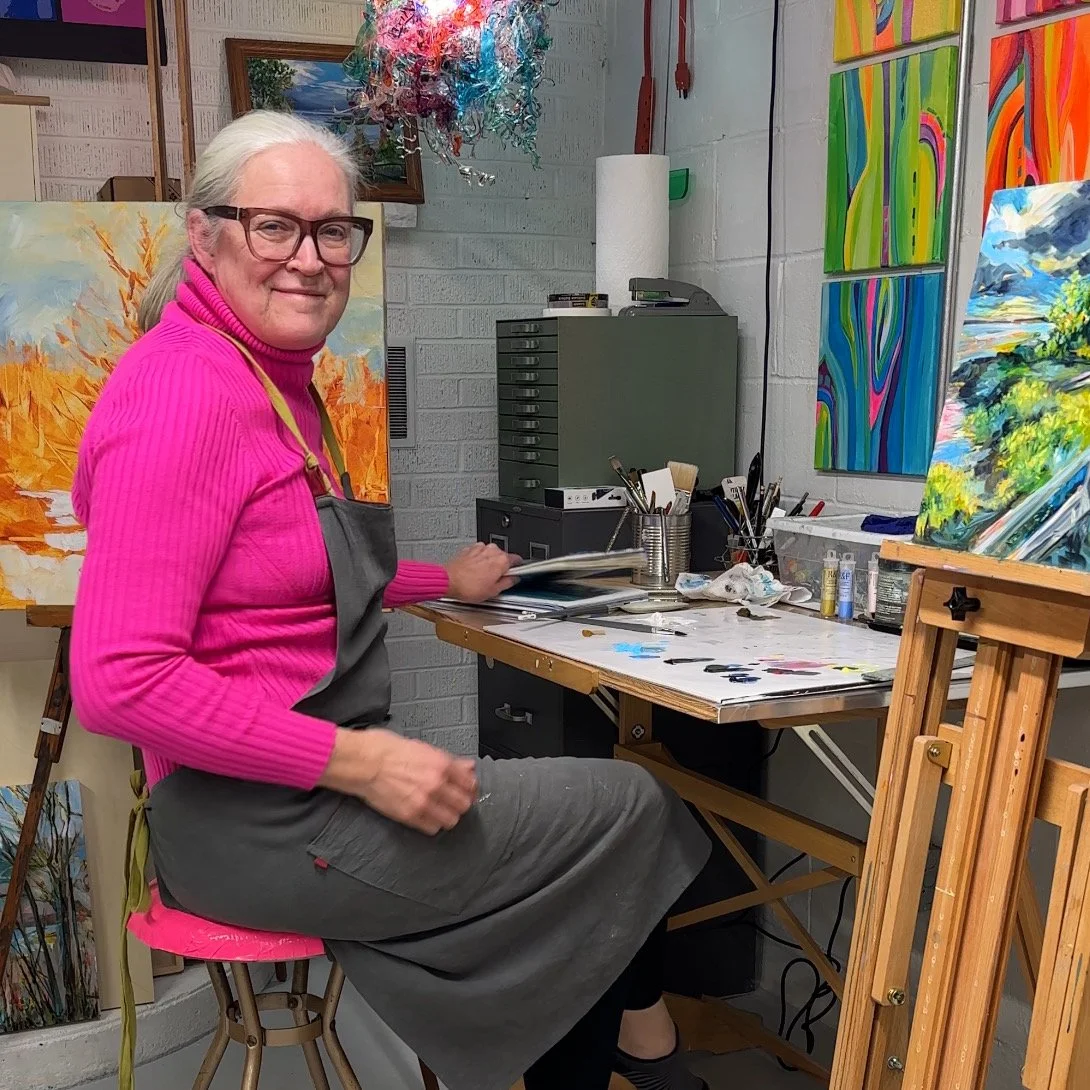 An older woman with gray hair, glasses, and a pink turtleneck sweater sitting on a stool in an art studio, surrounded by paintings and art supplies, smiling at the camera.