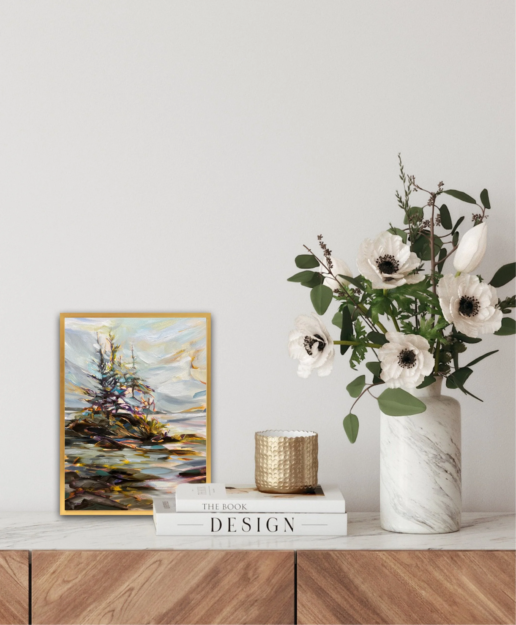 Decorative arrangement featuring a framed abstract painting, a white book titled 'Design,' a gold textured container, and a large white marble vase with white anemone flowers and greenery on a wooden surface against a plain wall.