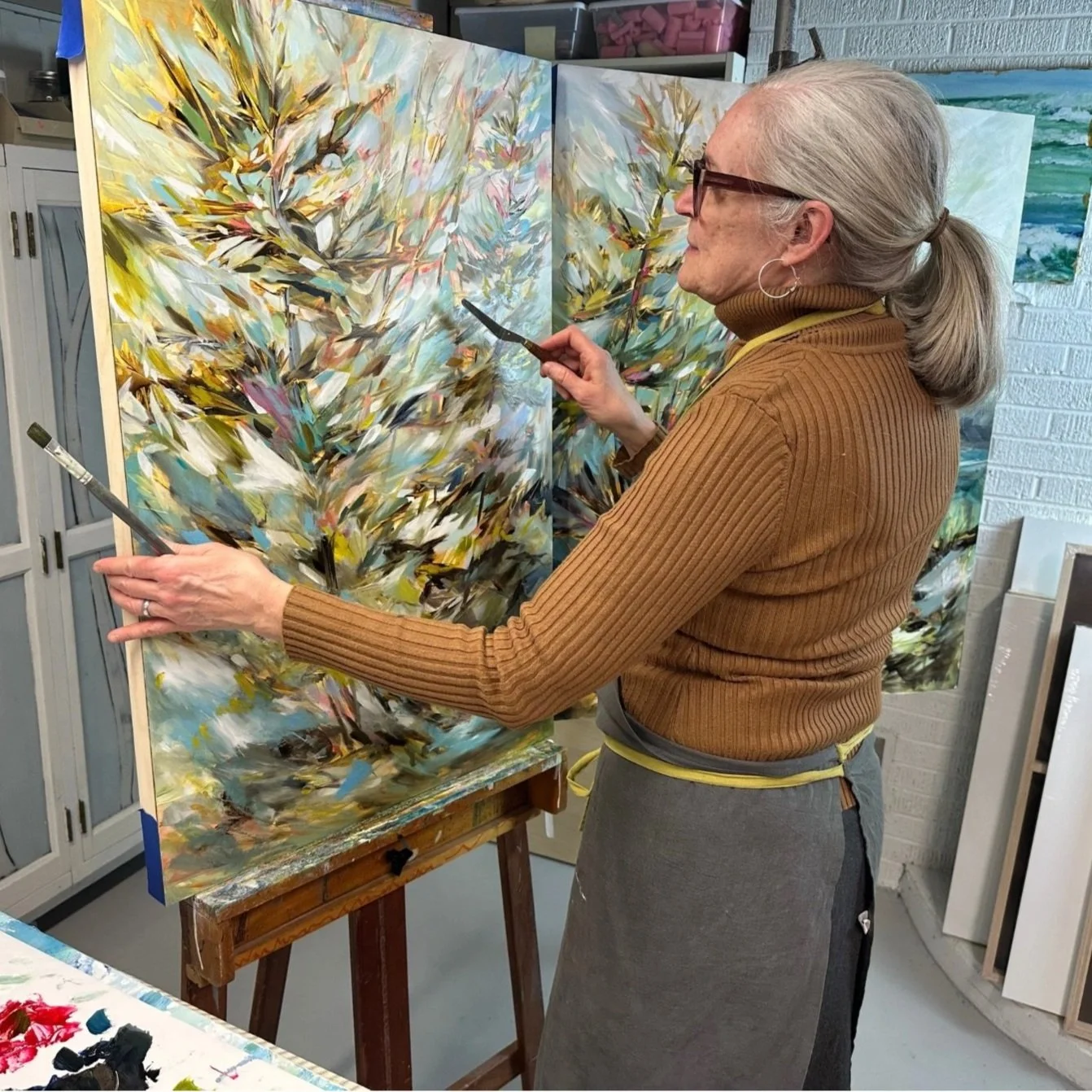 An artist in a brown turtleneck and gray apron painting an abstract landscape with bright, bold brushstrokes on a multi-panel canvas.