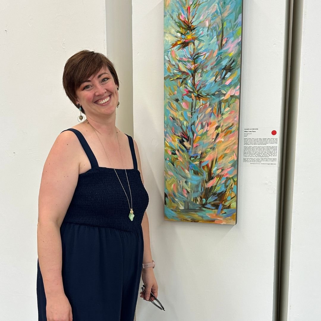 A woman with short brown hair, wearing a sleeveless black dress, smiling and standing next to an abstract colorful painting of a plant.