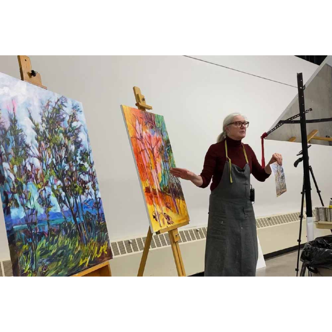An art instructor explaining her paintings on easels during a class. There are two colorful, abstract landscape paintings on display, one with a cloudy sky and the other with warm tones. The instructor is wearing glasses, a maroon turtleneck, and a gray apron, holding a small palette or cloth while gesturing with her other hand.