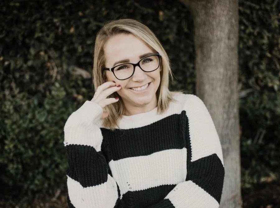 A woman with blonde hair and glasses smiling outdoors, wearing a black and white striped sweater with a background of greenery and a tree.