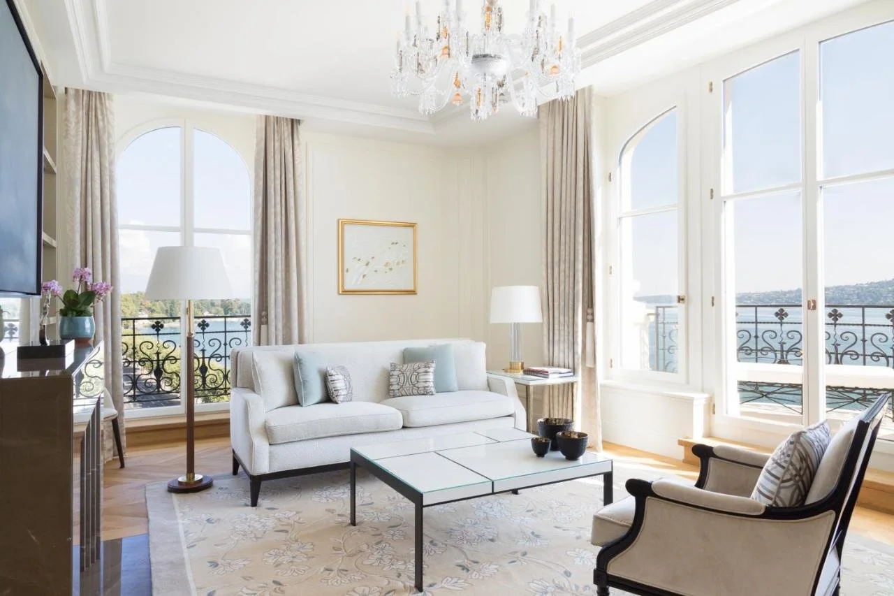 The Woodward Geneva grand Belle Époque suite living room with crystal chandelier and arched floor-to-ceiling windows with wrought iron balconies overlooking the blue water of Lake Geneva