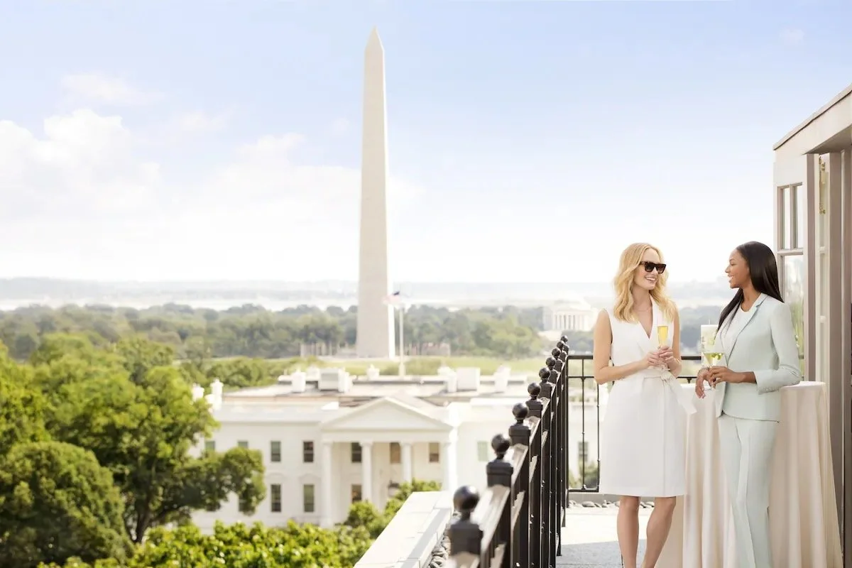 The Hay-Adams Washington DC rooftop terrace with two guests holding champagne flutes, the White House directly below and the Washington Monument rising above the treeline