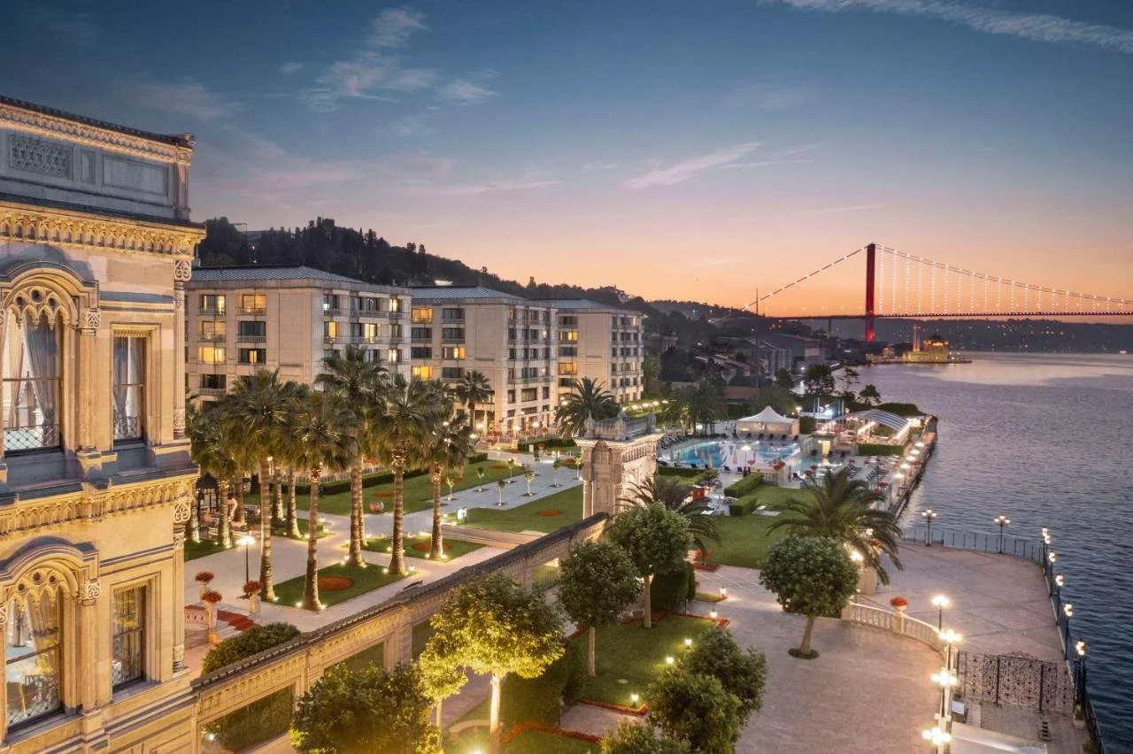 Çırağan Palace Kempinski Istanbul aerial view of the Ottoman palace facade and palm-lined gardens with the outdoor pool along the Bosphorus at sunset and the 15 July Martyrs Bridge in the background