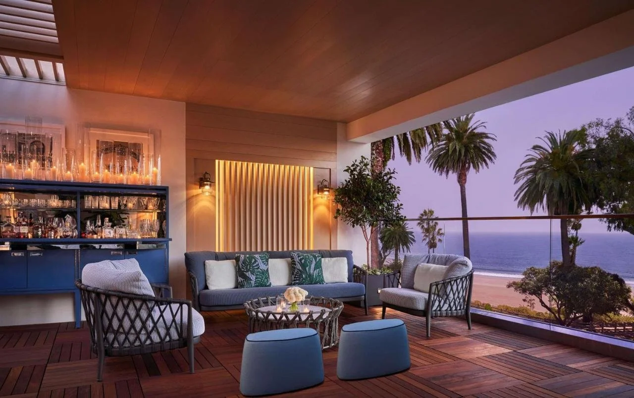 Oceana Santa Monica’s outdoor terrace lounge at dusk — teak decking, blue sofas, and a glass railing open to the Pacific and palm trees at golden hour