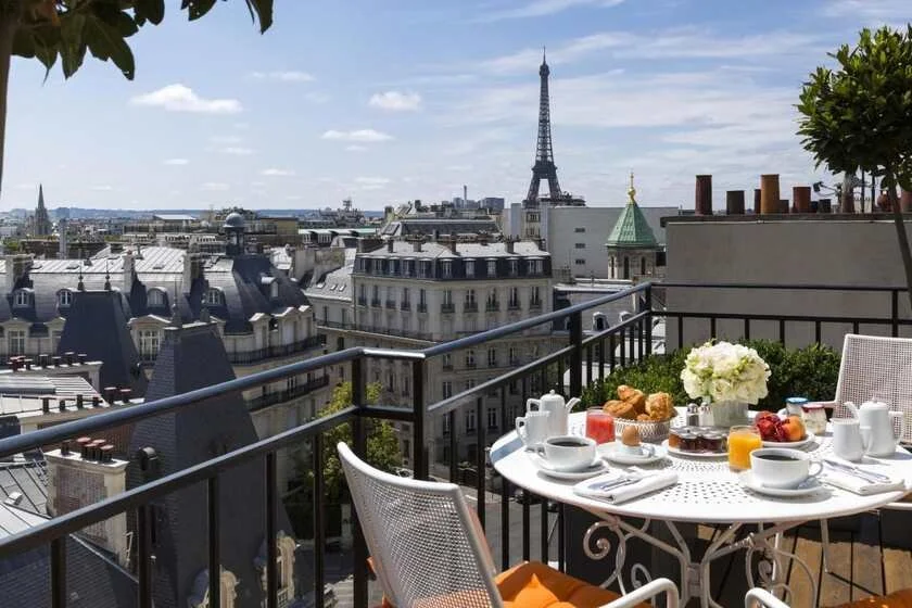 Hôtel San Régis Paris top-floor terrace breakfast with croissants, orange juice, and white roses, the Eiffel Tower rising above the 8th arrondissement rooftops and a green copper-domed church tower in the background
