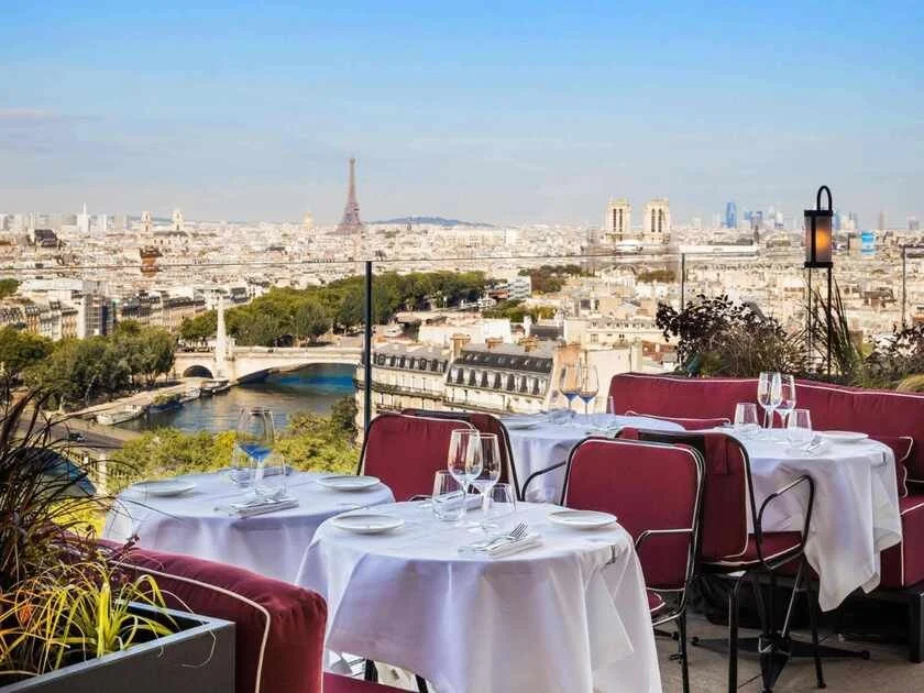 SO Paris Hotel rooftop restaurant terrace with white-linen tables and velvet red chairs, a panoramic view spanning the Seine, a bridge, the Eiffel Tower, Notre-Dame towers, and the La Défense skyline