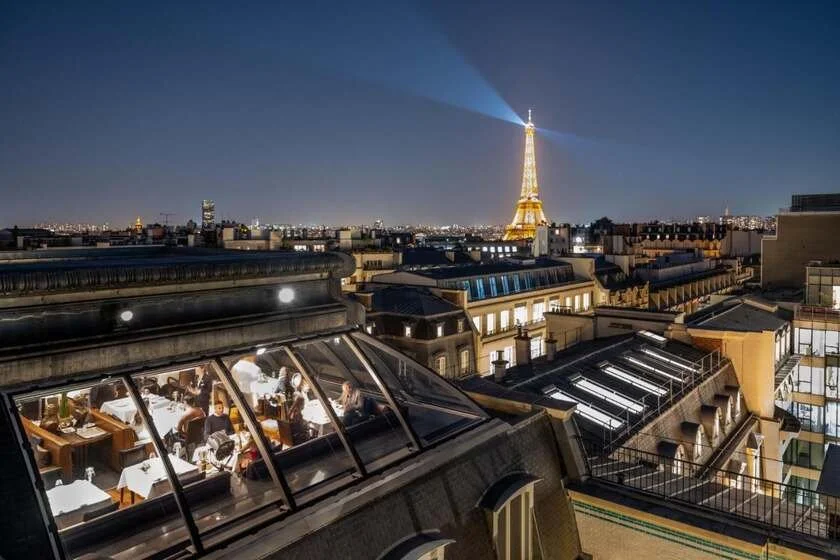 The Peninsula Paris night aerial view with a glass-roofed restaurant dining room visible below and the illuminated Eiffel Tower sending its rotating beacon light across the Paris skyline