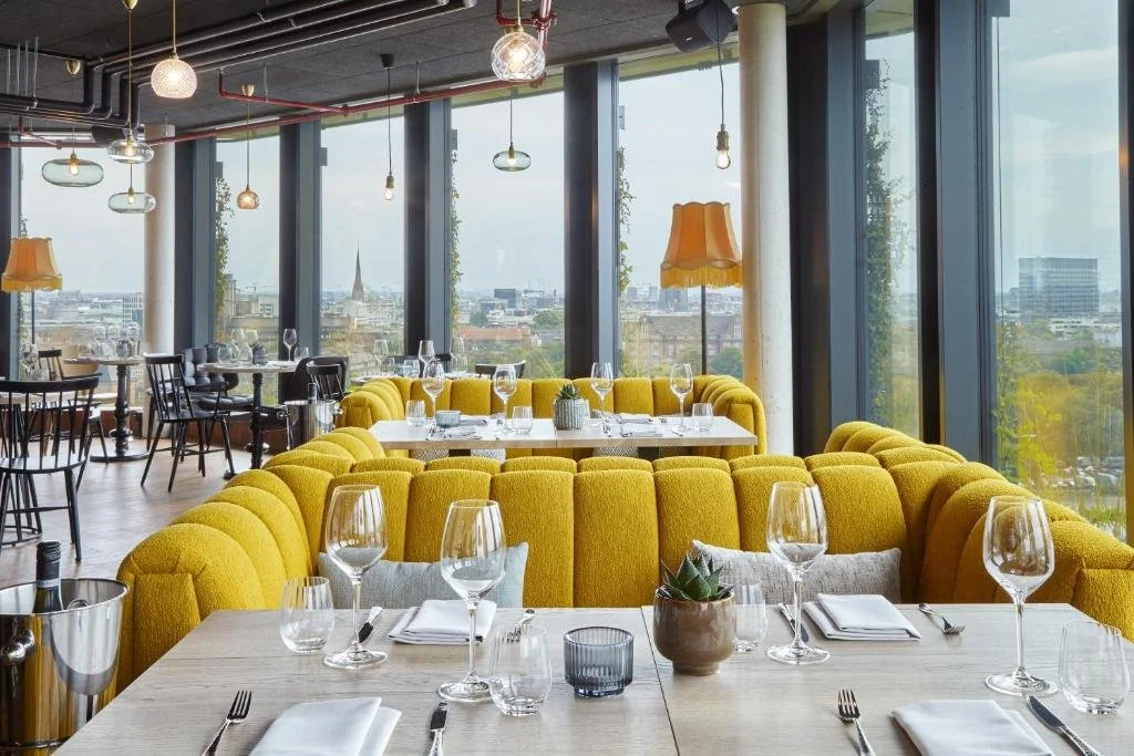 REVERB by Hard Rock Hamburg rooftop restaurant with mustard yellow curved booth seating and floor-to-ceiling windows overlooking Hamburg rooftops and a church spire