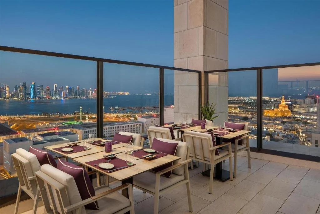 Park Hyatt Doha Doha Sora rooftop restaurant terrace at dusk with set tables and panoramic view of the illuminated West Bay skyline, the Arabian Gulf, and the Fanar Islamic Cultural Center spiral minaret