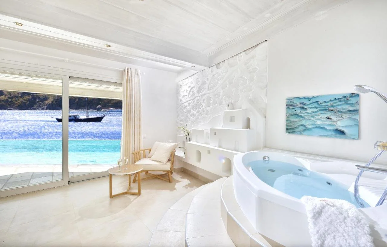 Nissaki Boutique Hotel Mykonos all-white bathroom with jacuzzi tub and glass door opening to pool terrace and Aegean Sea with traditional boat