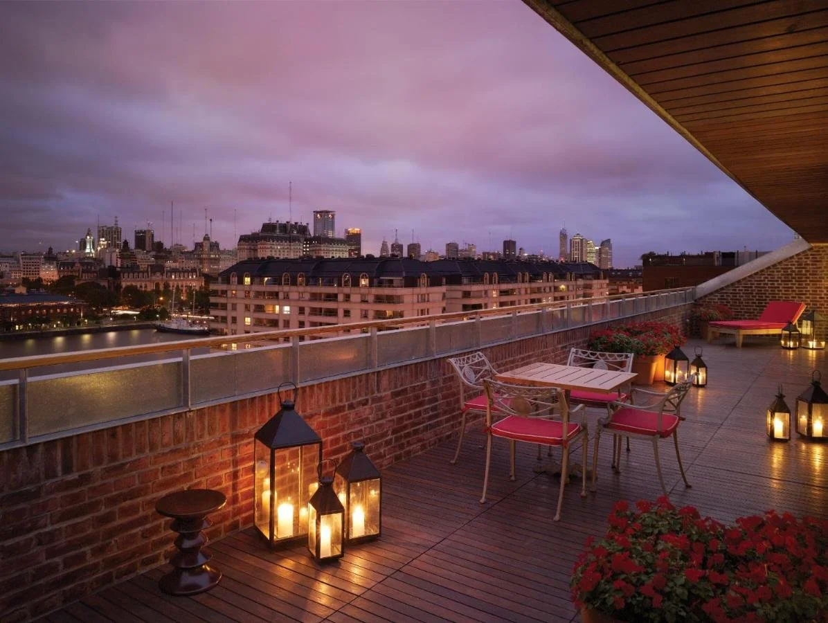 Faena Hotel Buenos Aires rooftop terrace at dusk with wrought-iron garden furniture, lanterns, red geraniums, and waterfront skyline under purple sky