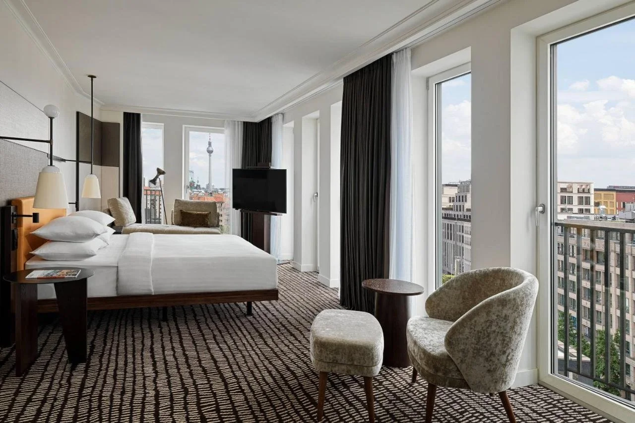 Berlin Marriott Hotel Berlin elegant hotel room with upholstered seating, dark curtains and three tall windows with the Berlin TV Tower visible in the distance and a balcony overlooking Mitte rooftops