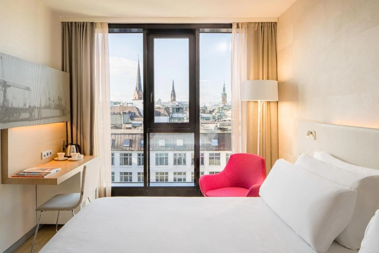 Barceló Hamburg hotel room with white bed and red armchair facing a floor-to-ceiling window with views of multiple Hamburg church spires rising above the city rooftops