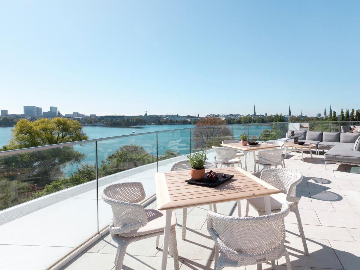 The Fontenay Hamburg rooftop terrace with white lattice chairs and wooden tables overlooking the Außenalster Lake, Hamburg church spires visible across the water on the far shore