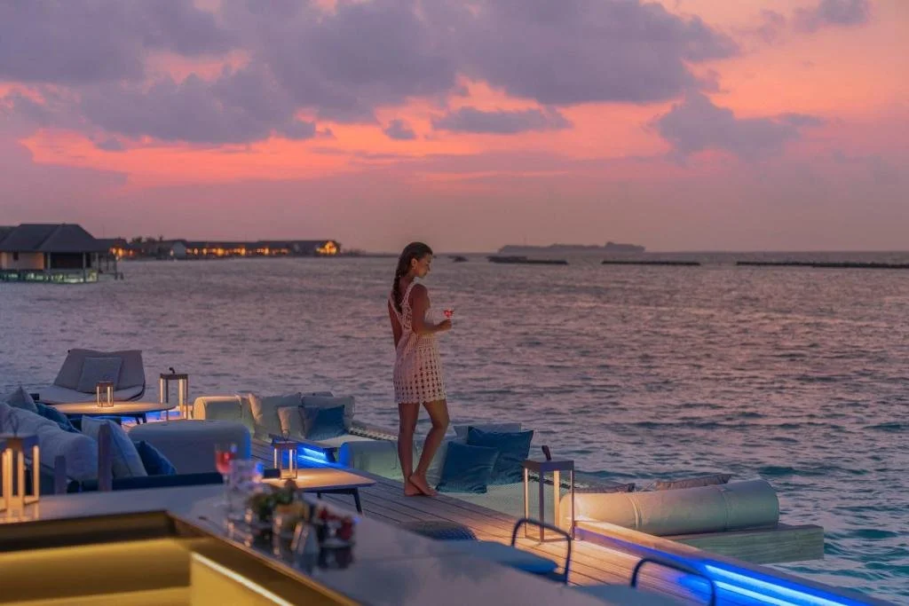 Ritz-Carlton Maldives Fari Islands overwater villa infinity pool at ocean edge with two guests wading, circular day bed and wicker table on teak deck